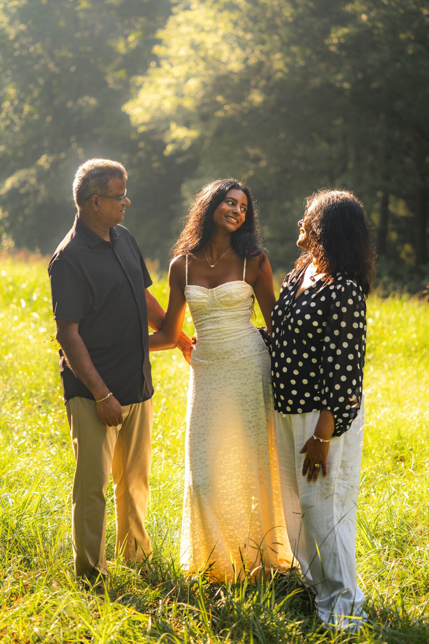 A young woman in a white dress stands with two older adults in a sunlit green field.