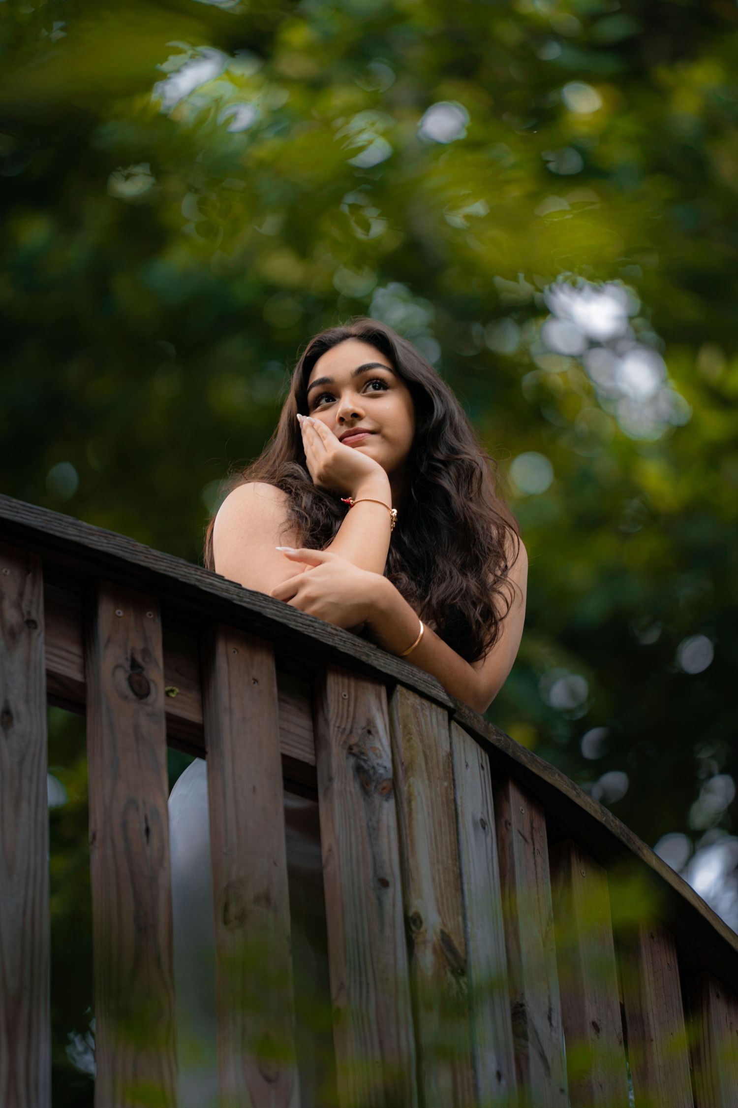 Young woman with long dark hair leans on wooden bridge railing, gazing upward dreamily, surrounded by lush green trees.