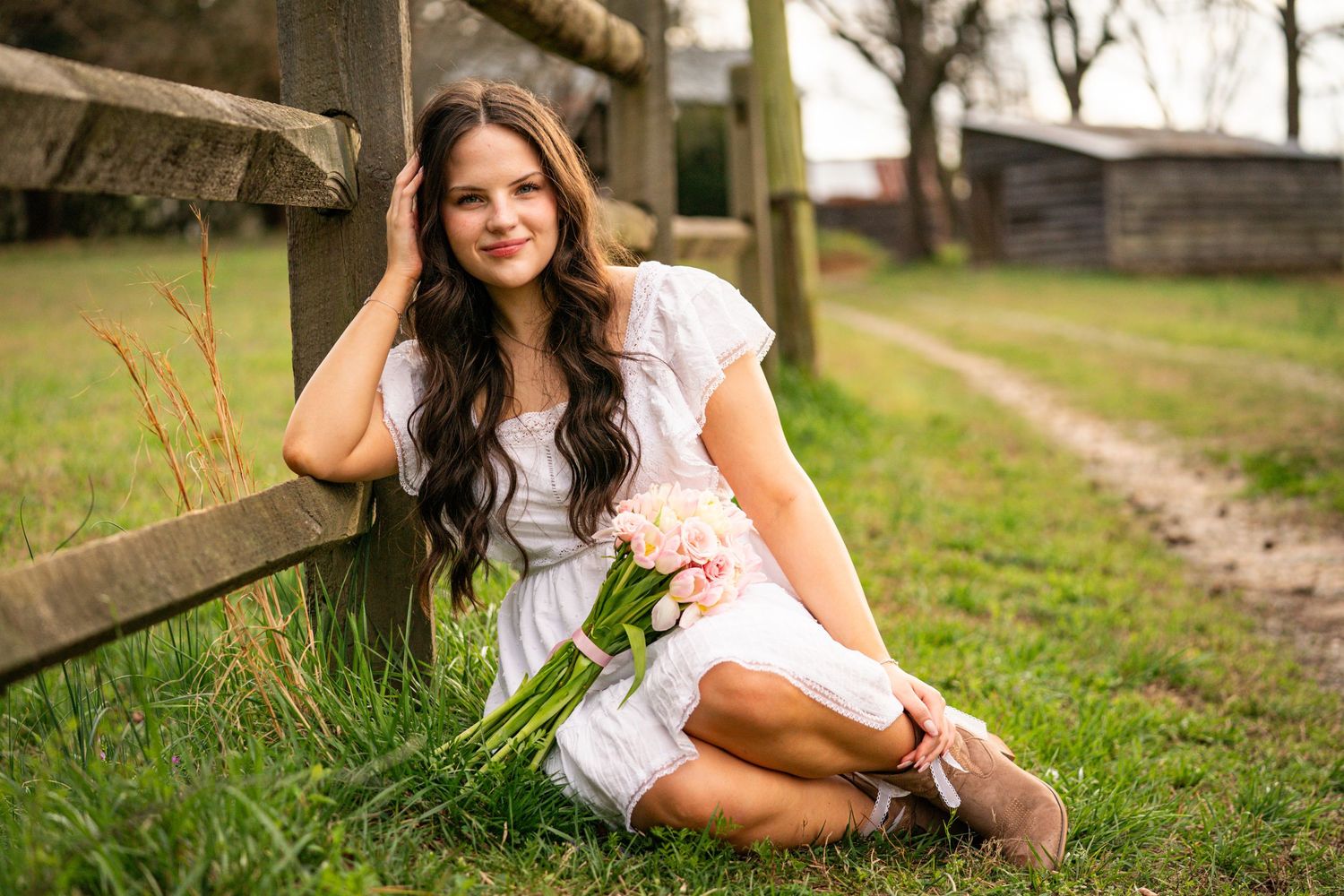 A woman in a white dress sits gracefully on grass near a rustic wooden fence in an outdoor setting.