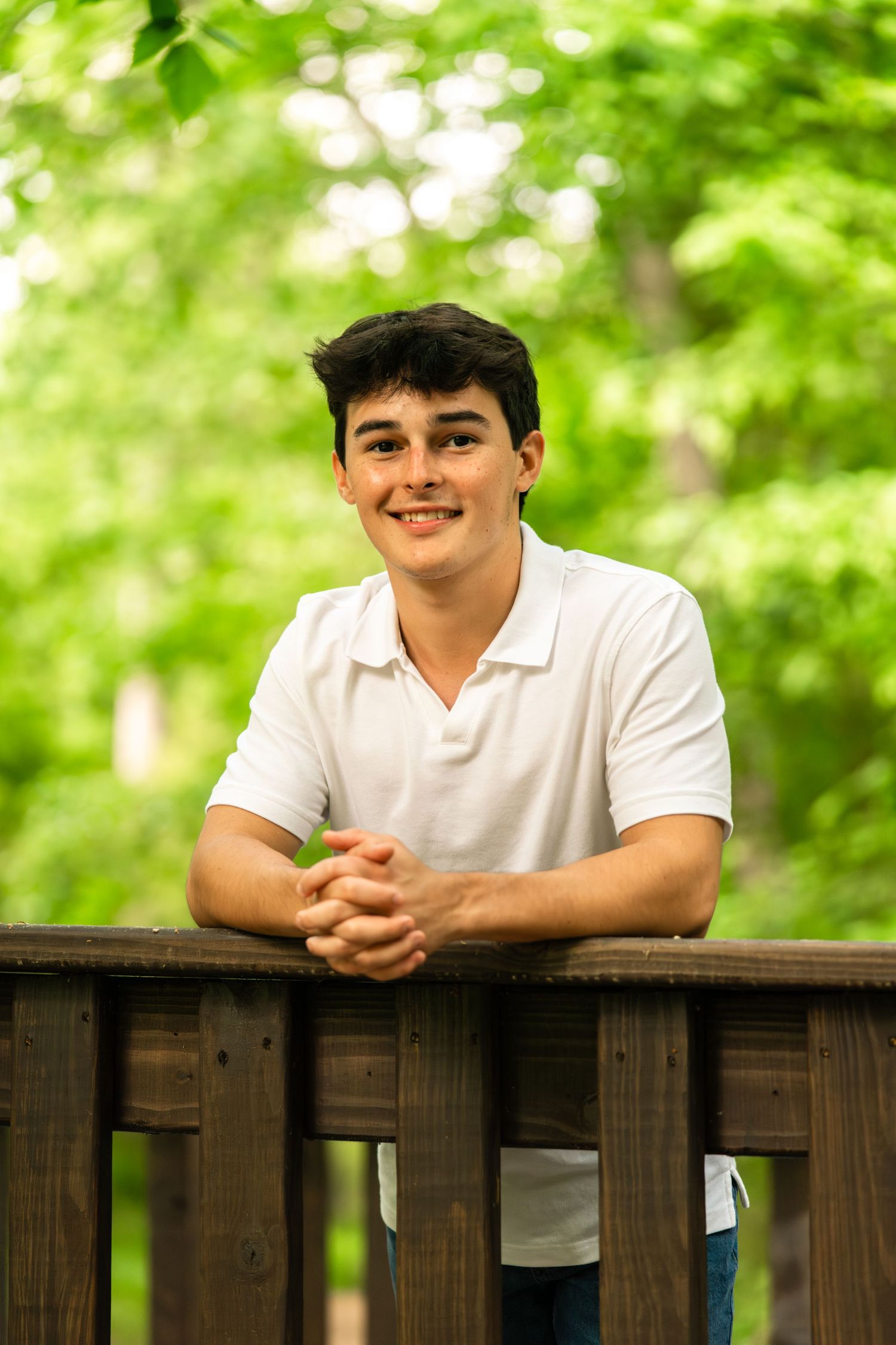 Smiling young man in white polo shirt leaning on wooden railing outdoors with lush green trees in background.