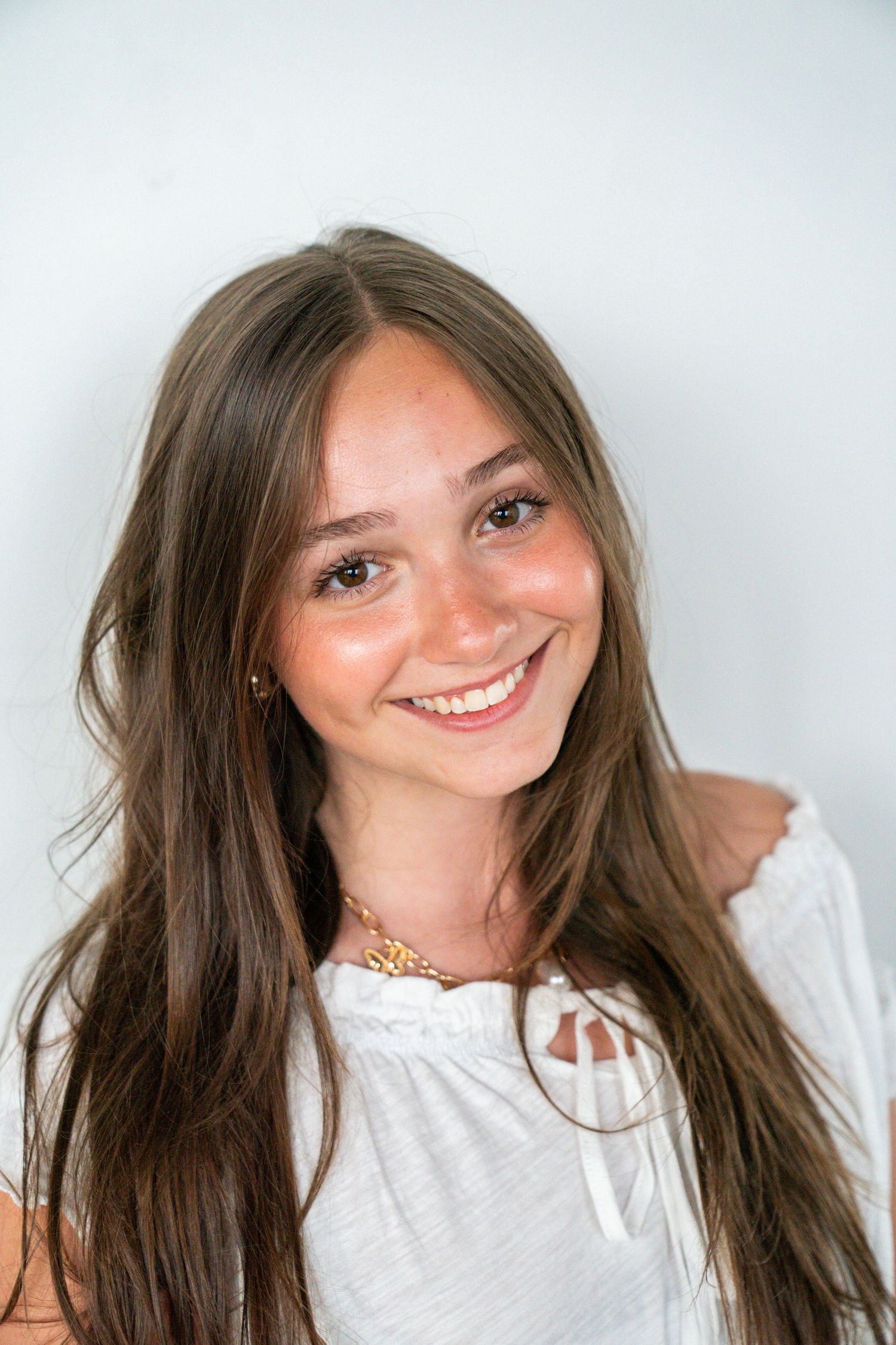 Smiling young woman with long brown hair wearing a white top and gold necklace against a light background.