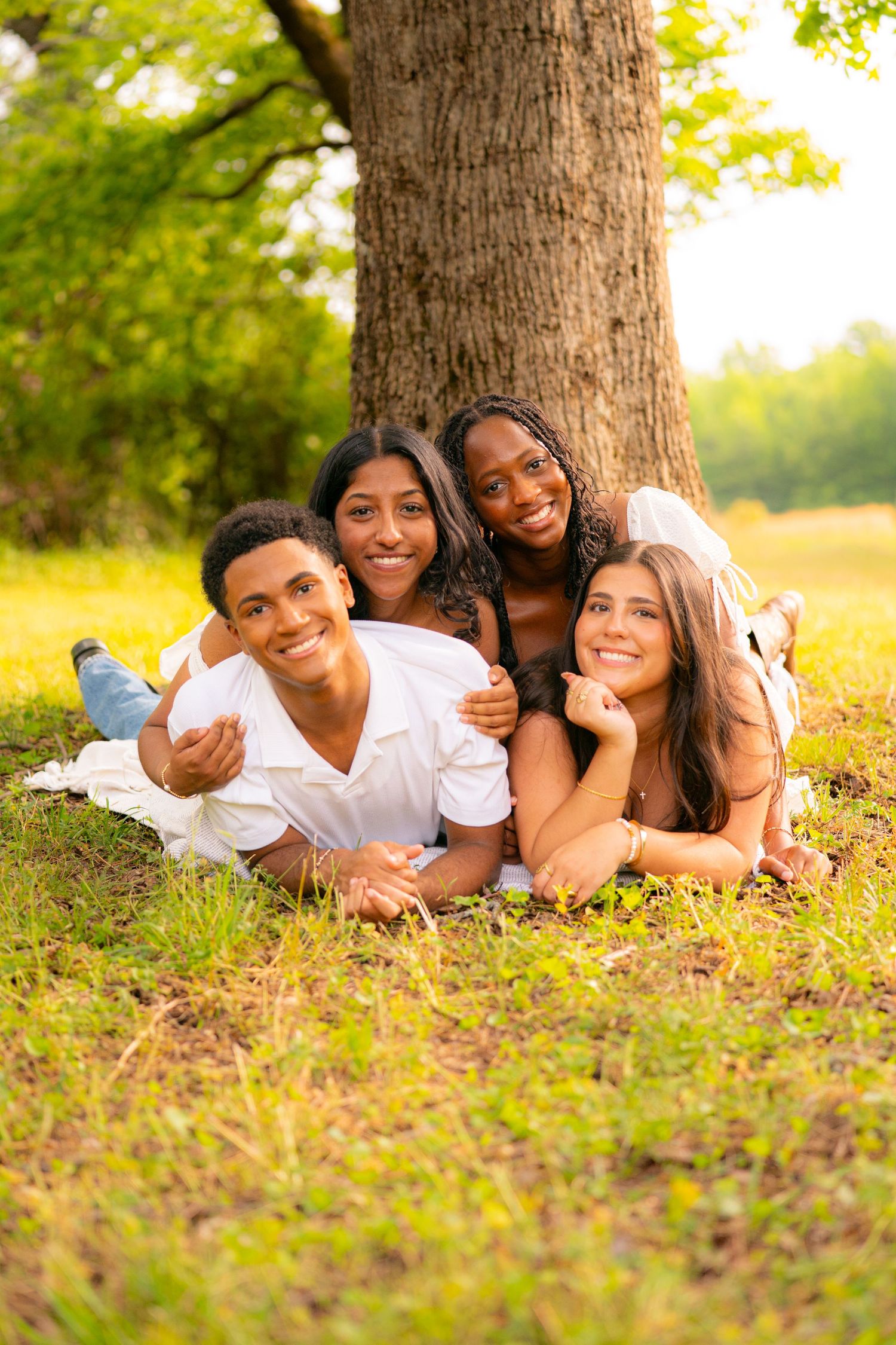 Four diverse smiling teenagers lying on grass under a tree in a sunny park, enjoying a fun outdoor day together.