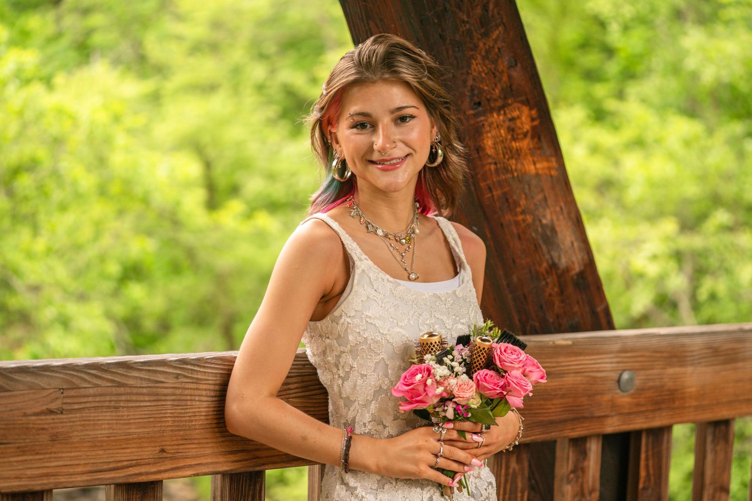 Young woman in white dress holding pink roses bouquet, smiling on wooden bridge surrounded by green trees.