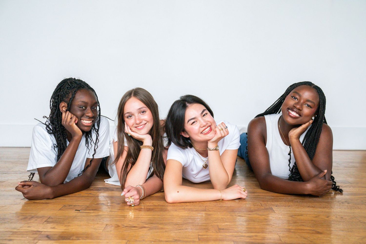 Four diverse young girls smiling and lying on a wooden floor against a white wall.
