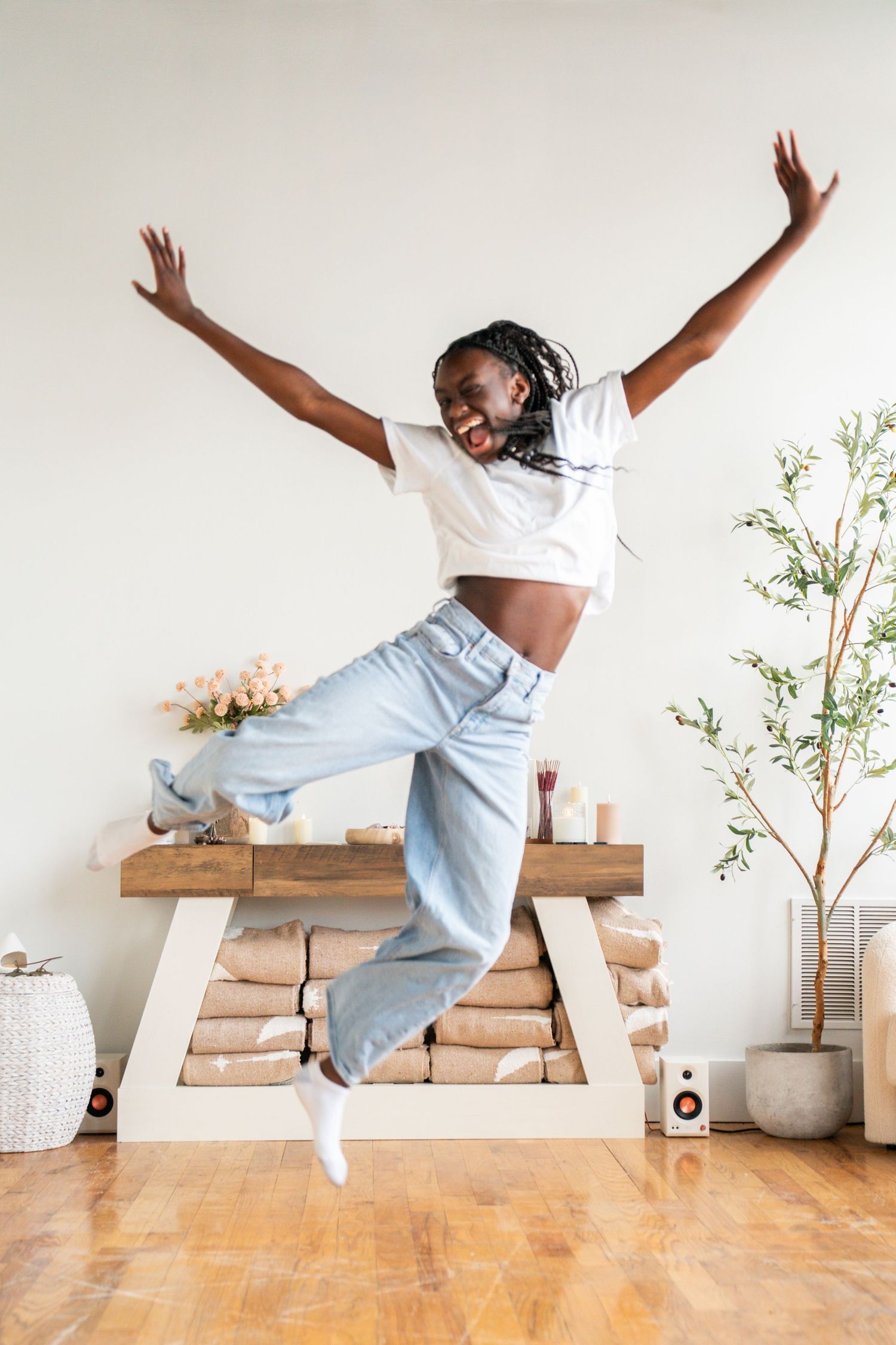 Joyful young Black woman jumping with arms raised in bright living room, wearing white crop top and light blue jeans.
