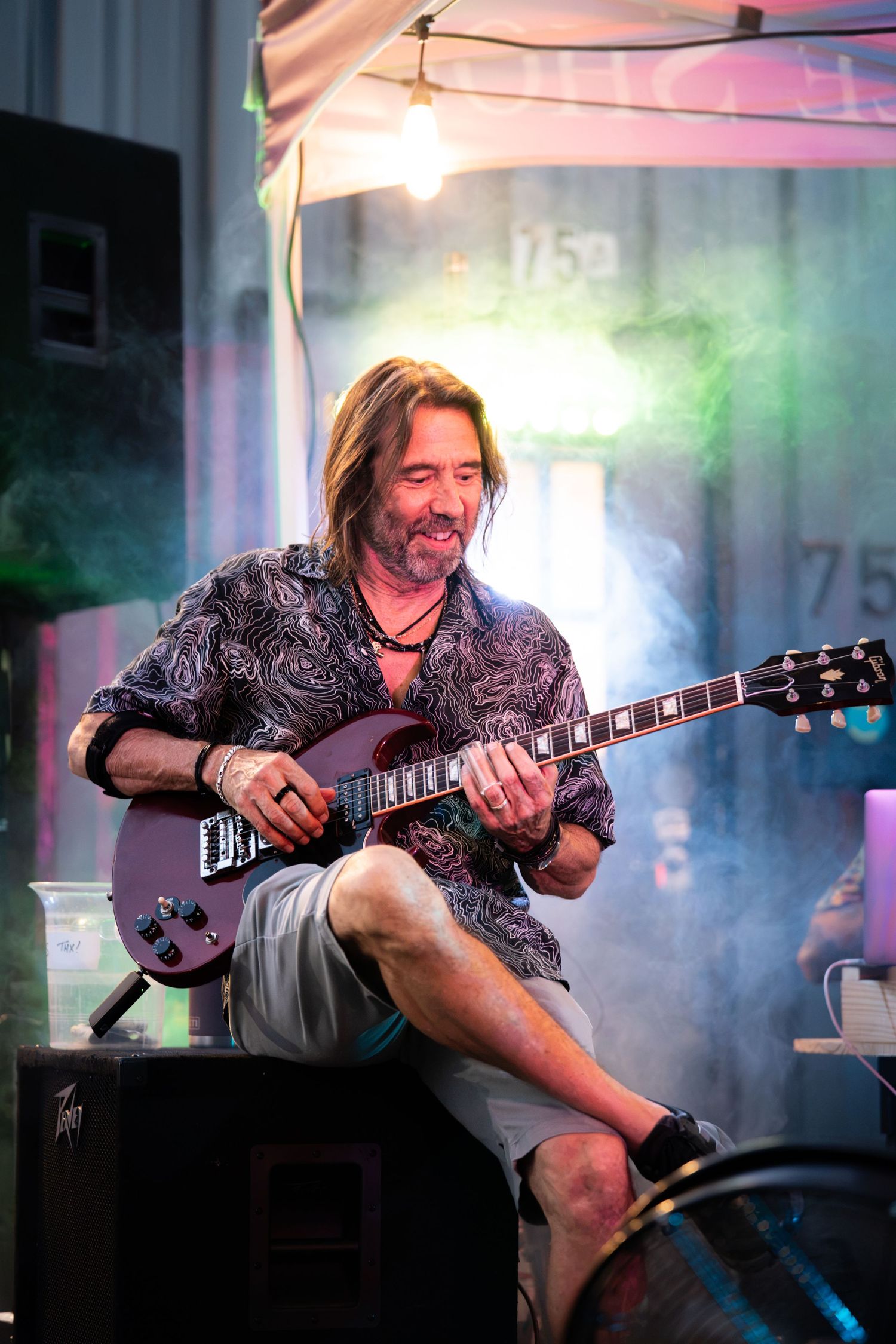 A smiling man with long hair plays an electric guitar while sitting on an amplifier at an outdoor music performance.