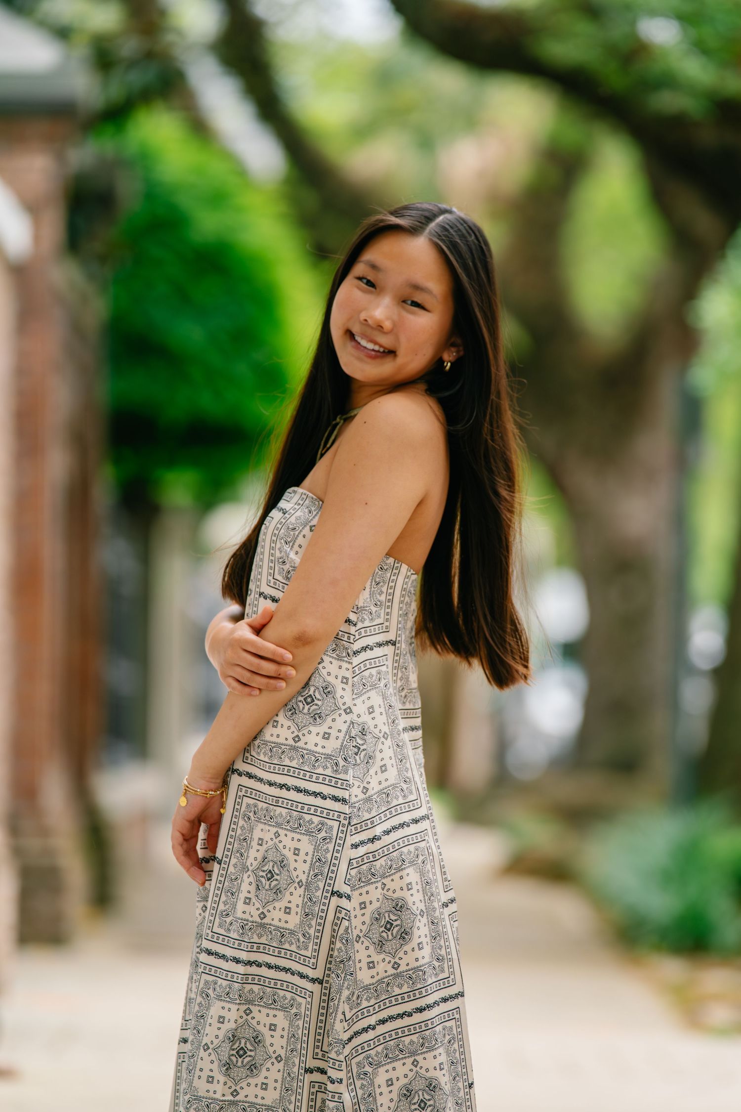 Young Asian woman smiling in patterned sundress on tree-lined brick pathway outdoors.