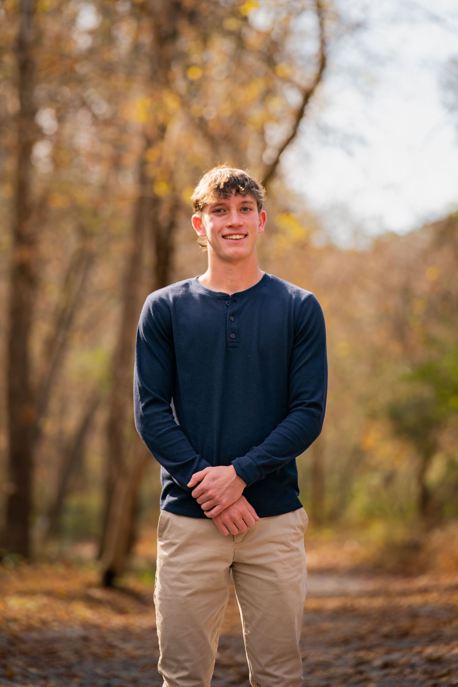 Student in navy henley and khakis stands on autumn forest path.