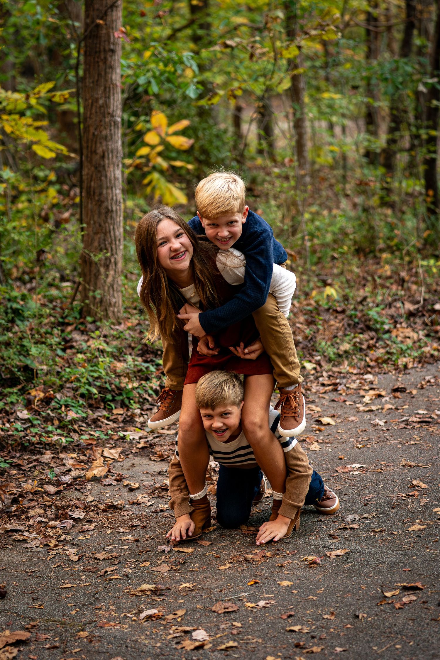 Playful outdoor photoshoot showing piggyback rides and laughter on a forest path covered in fallen leaves.
