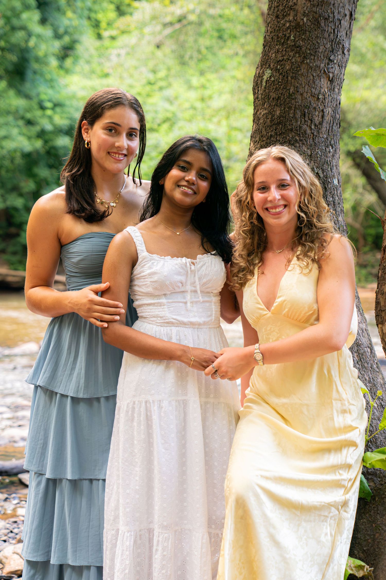 Three young women in elegant dresses posing together outdoors near a creek surrounded by lush green trees.