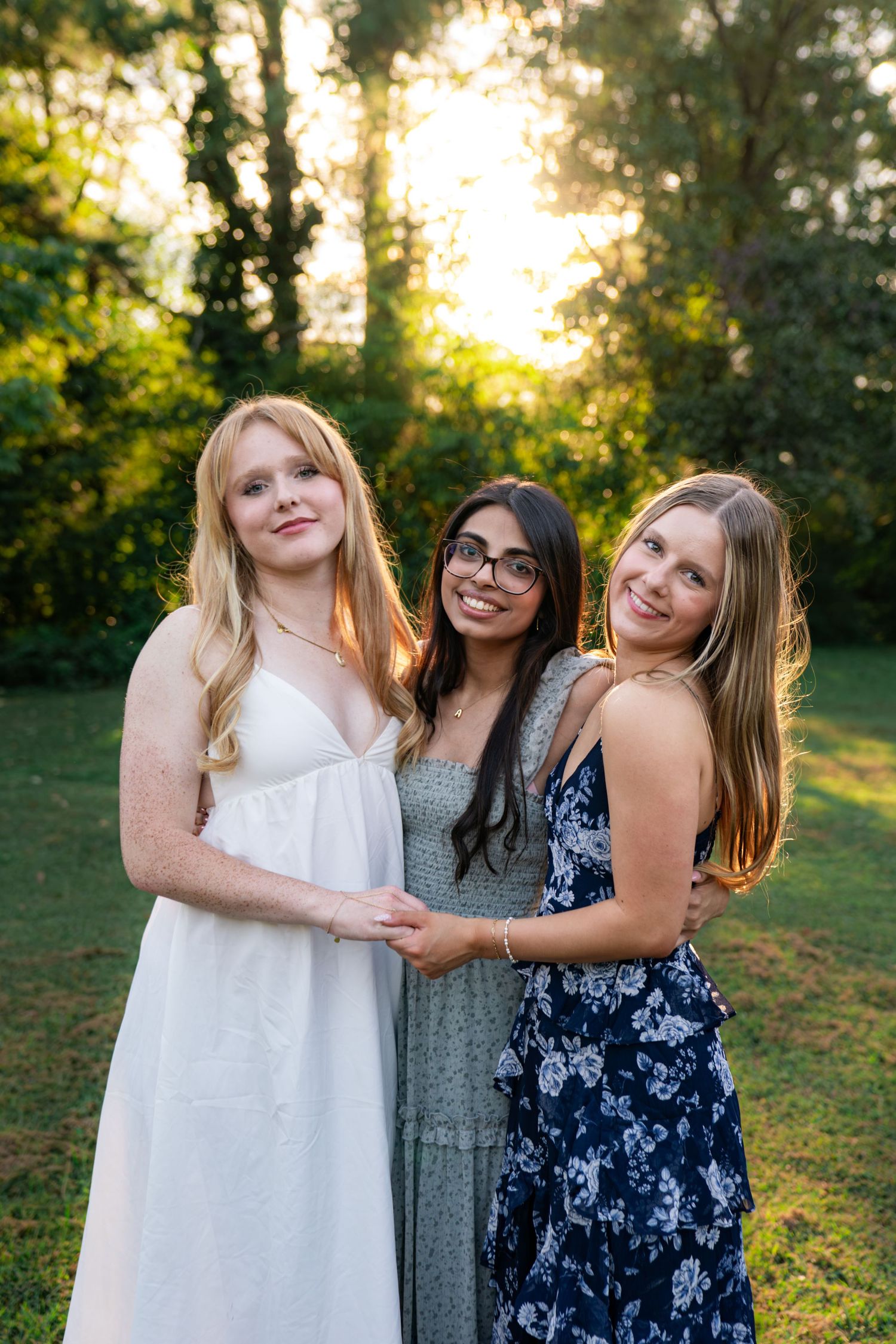 Three smiling young women in summer dresses embrace outdoors on a grassy lawn with golden sunset light filtering through green trees.