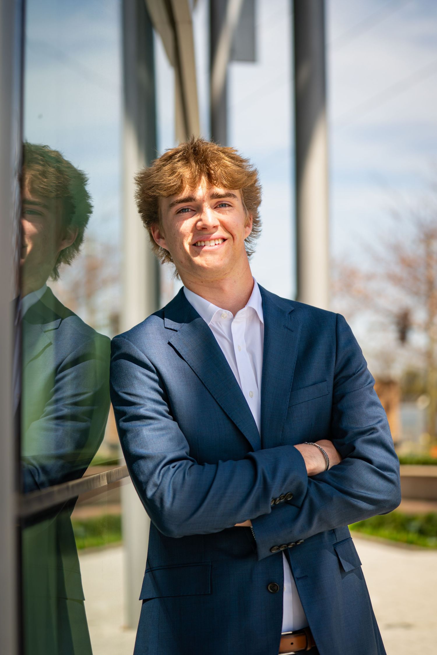 Professional in a navy blue suit and white shirt leans against a glass wall with reflections in an outdoor setting.