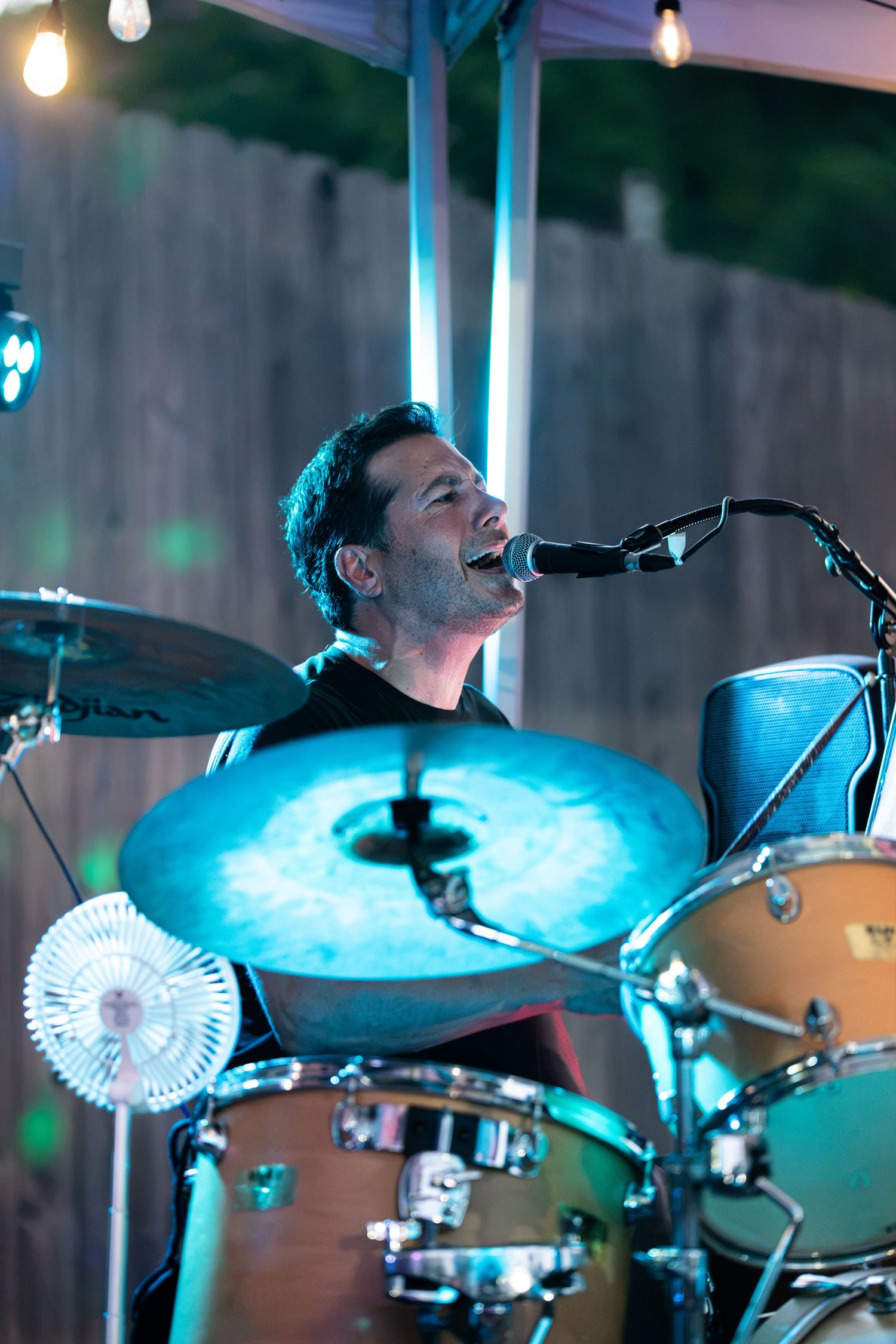 Male drummer singing into microphone while playing drums on an outdoor stage with colorful lighting.