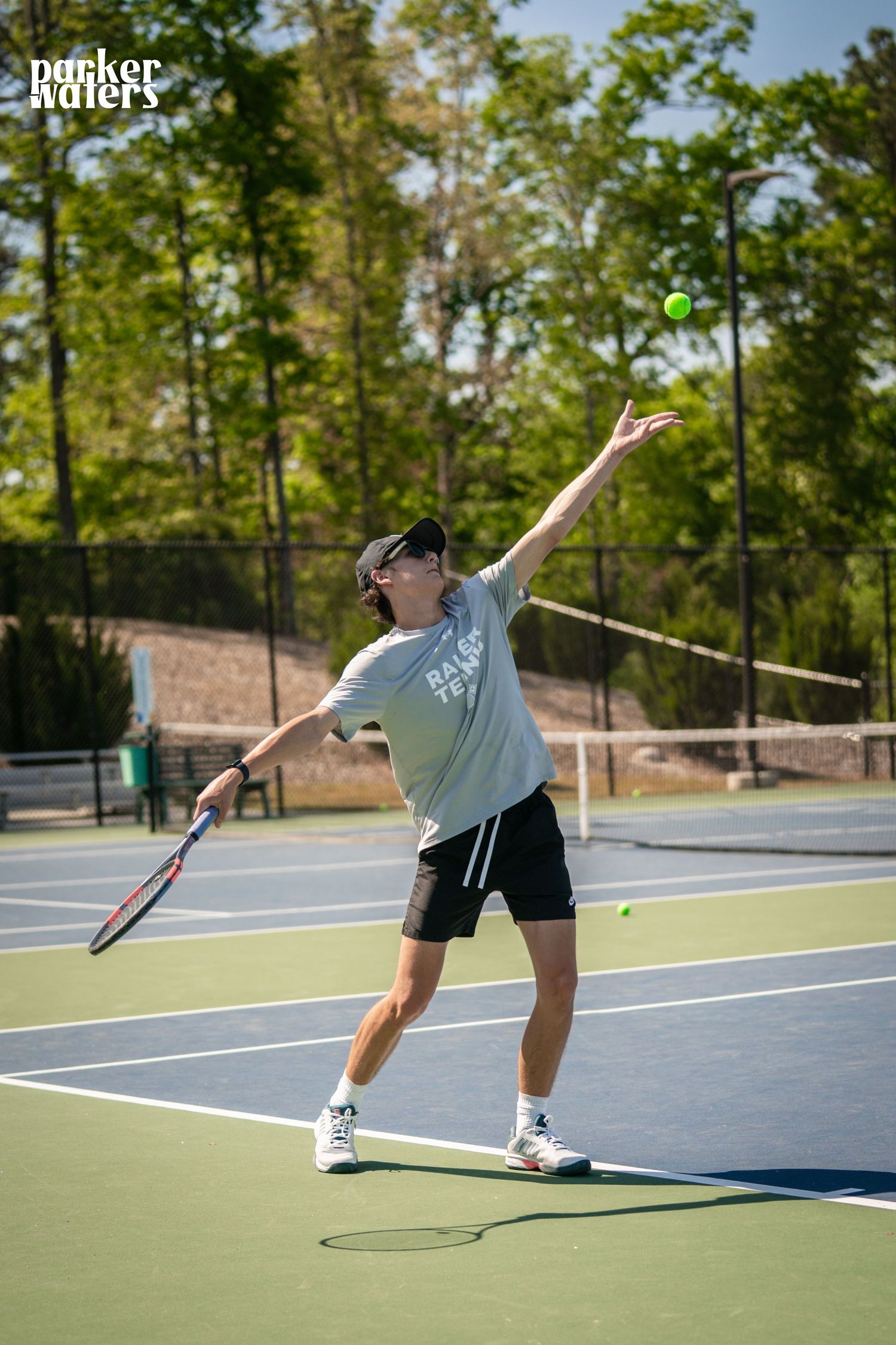 A man serves a tennis ball on an outdoor court, wearing a gray shirt, black shorts, and sunglasses on a sunny day.