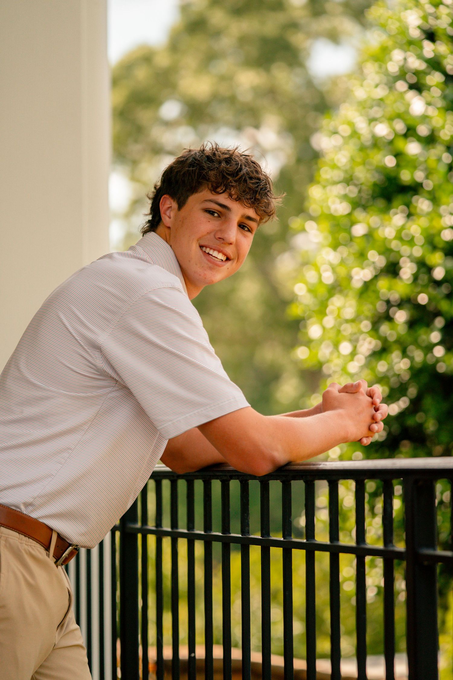 Smiling young man in white polo shirt leaning on black railing with lush green trees in background.