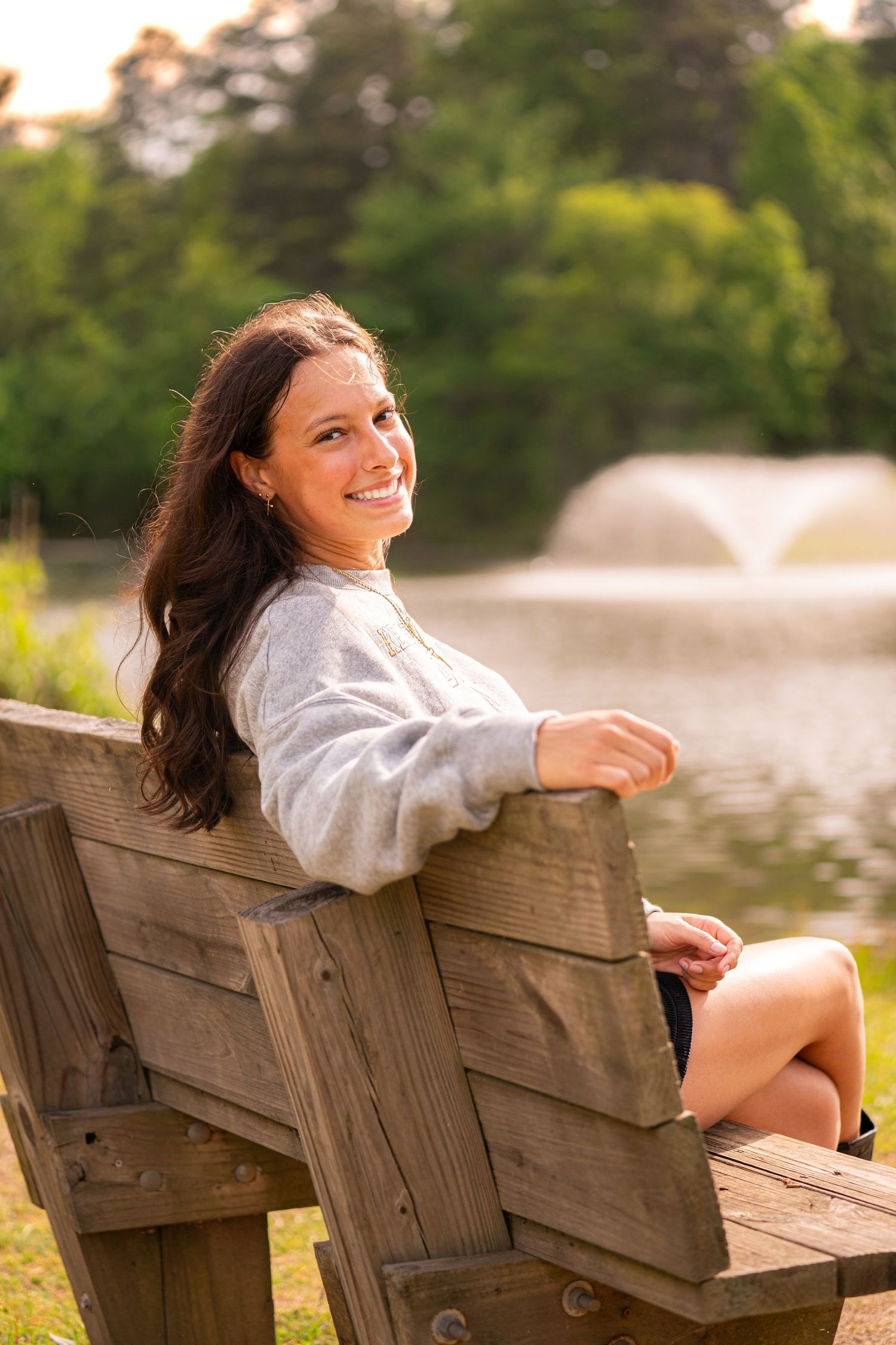 Young woman with long dark hair smiling on a wooden park bench near a pond with a fountain.