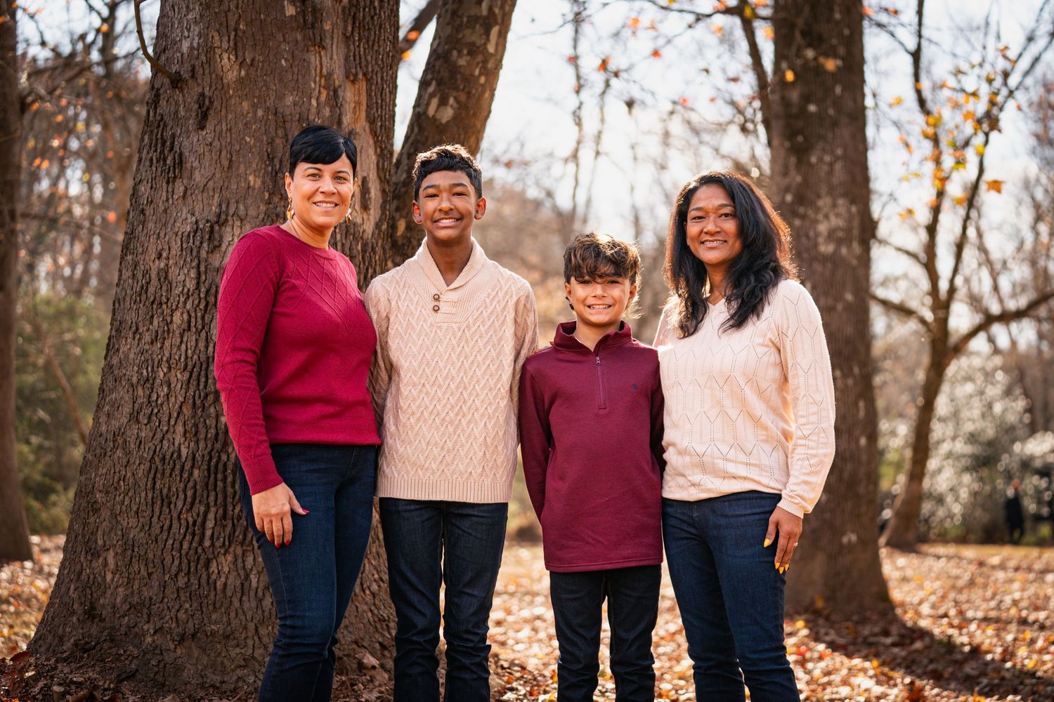 A family of four wearing cozy sweaters poses together in front of large trees during autumn.