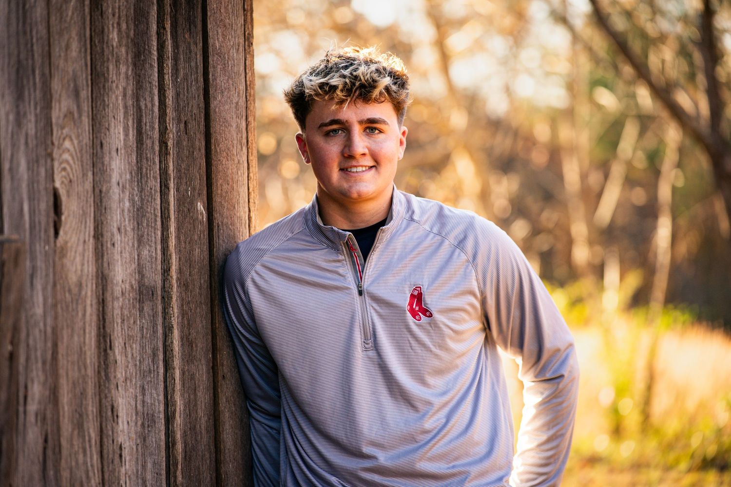 Person in a gray pullover smiles during golden hour outdoor photoshoot against wooden fence background.