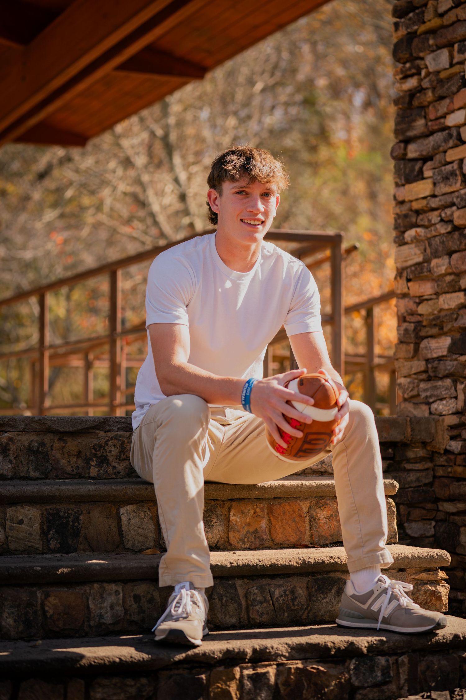 Student sits on stone steps holding football in outdoor autumn setting.