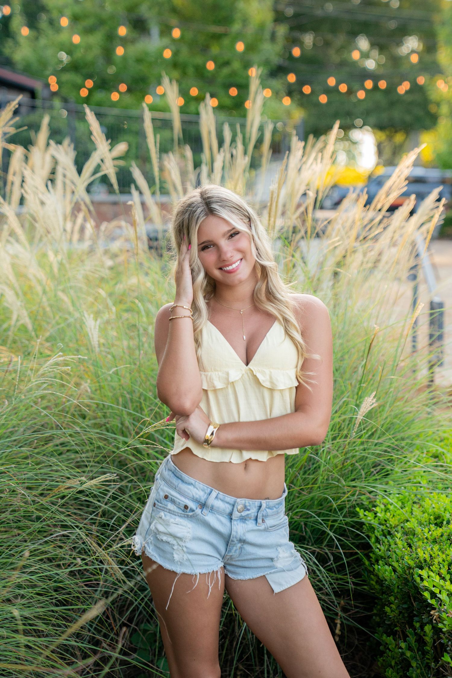 Smiling blonde woman in yellow crop top and denim shorts posing among tall ornamental grasses with string lights.