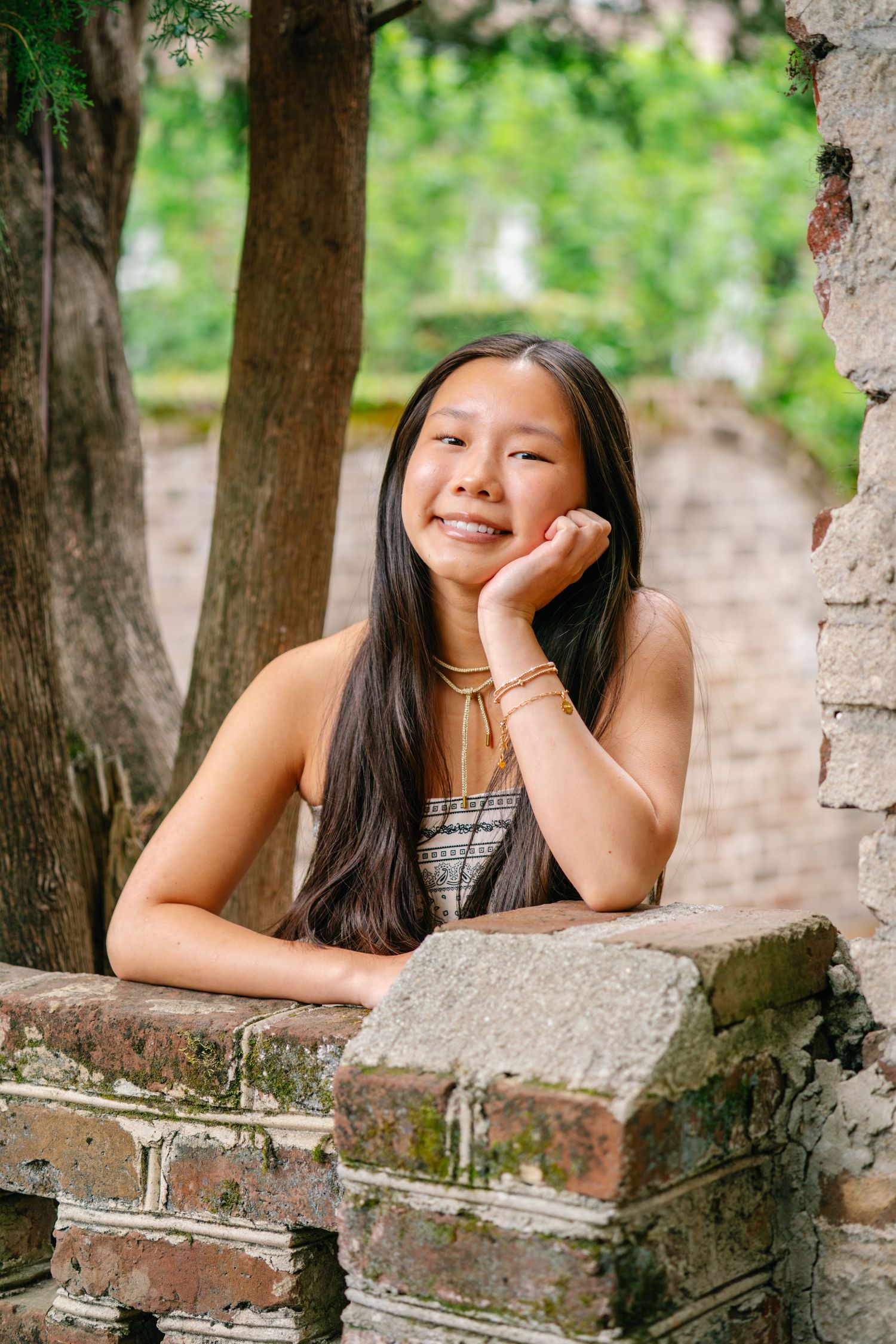 Young Asian woman smiling outdoors, leaning on mossy brick wall surrounded by lush green trees.