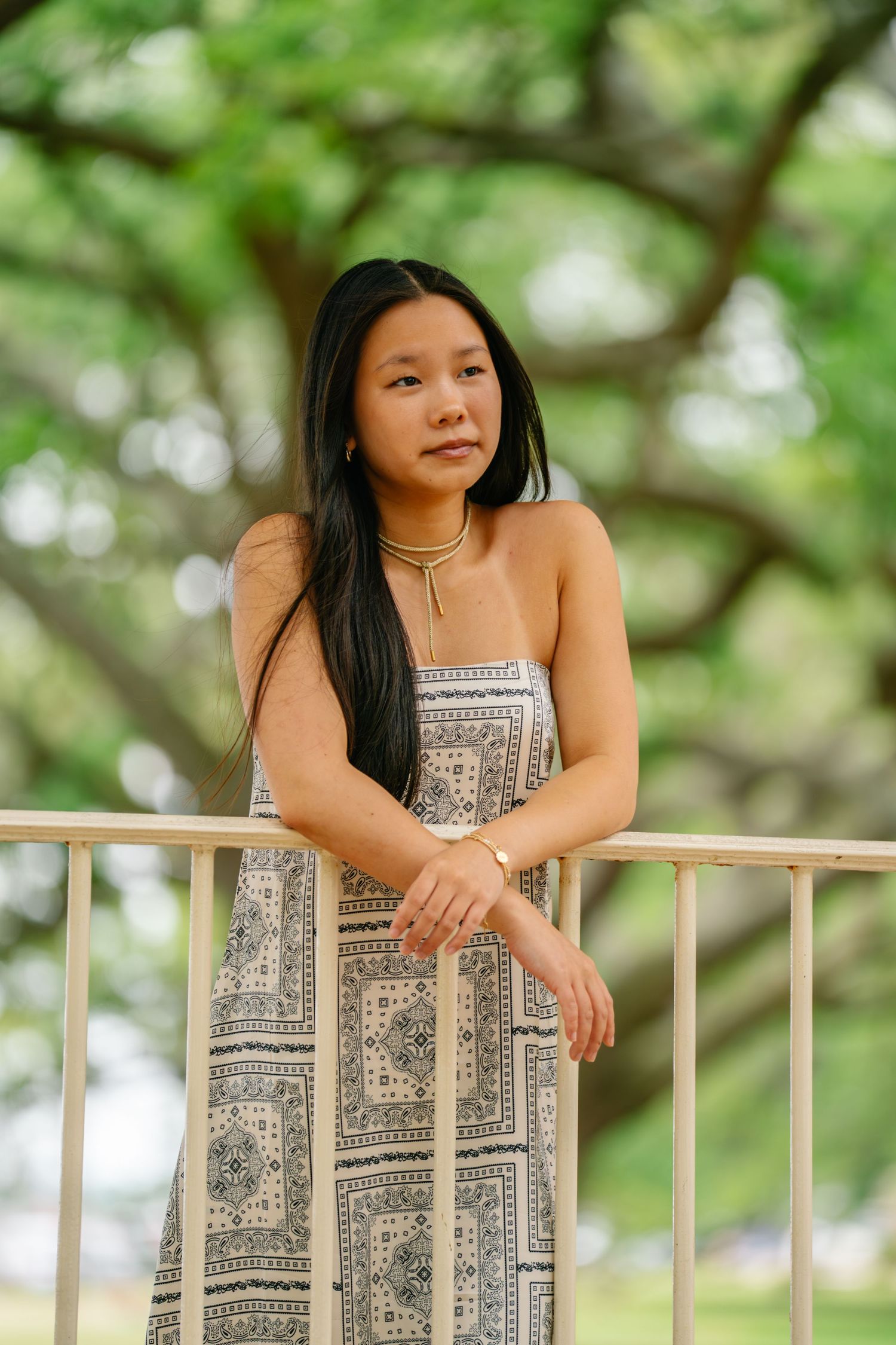 Young Asian woman in patterned sundress leaning on white railing outdoors with lush green trees in background.