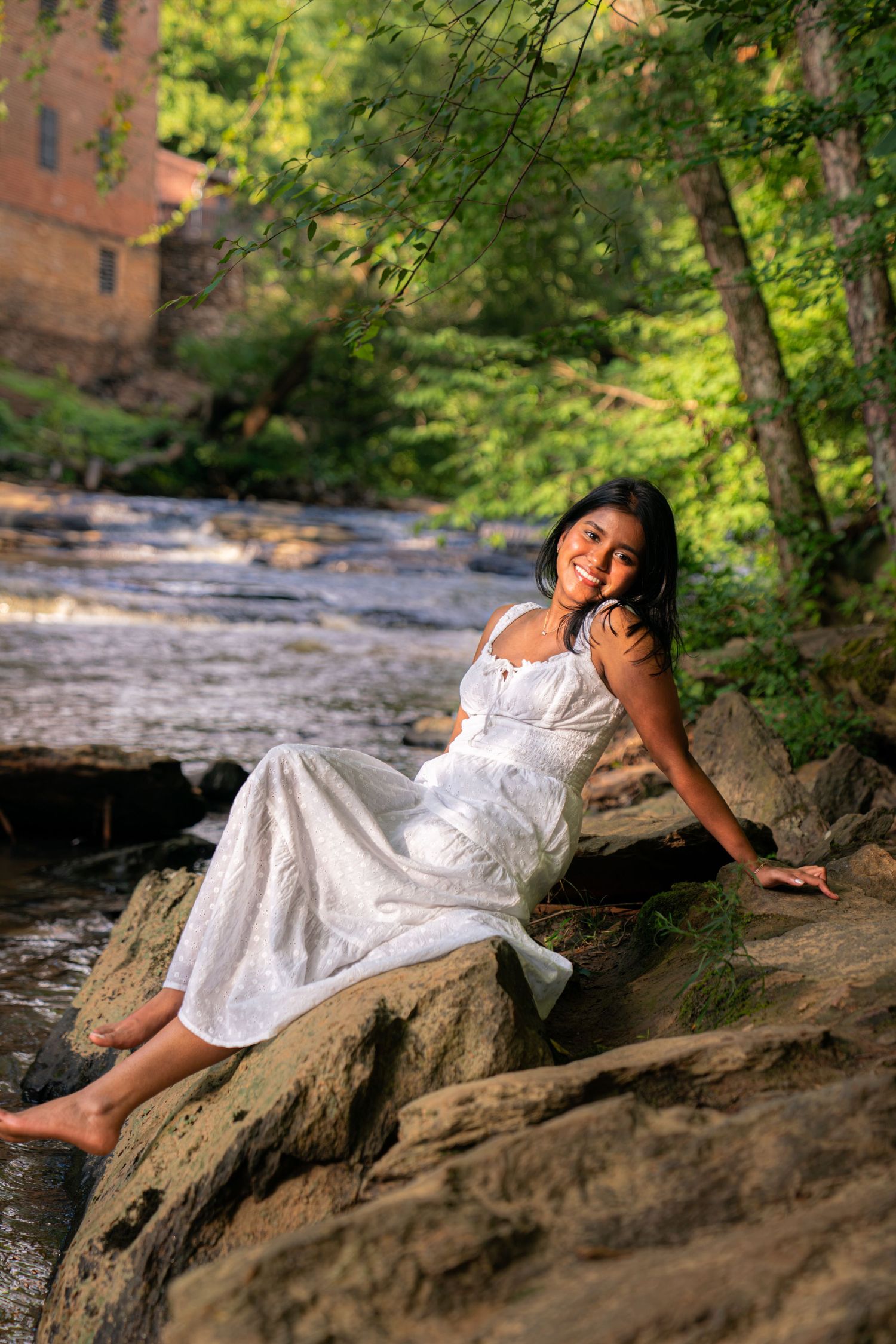 A smiling woman in a white dress sits barefoot on rocks beside a flowing stream in a lush green forest.