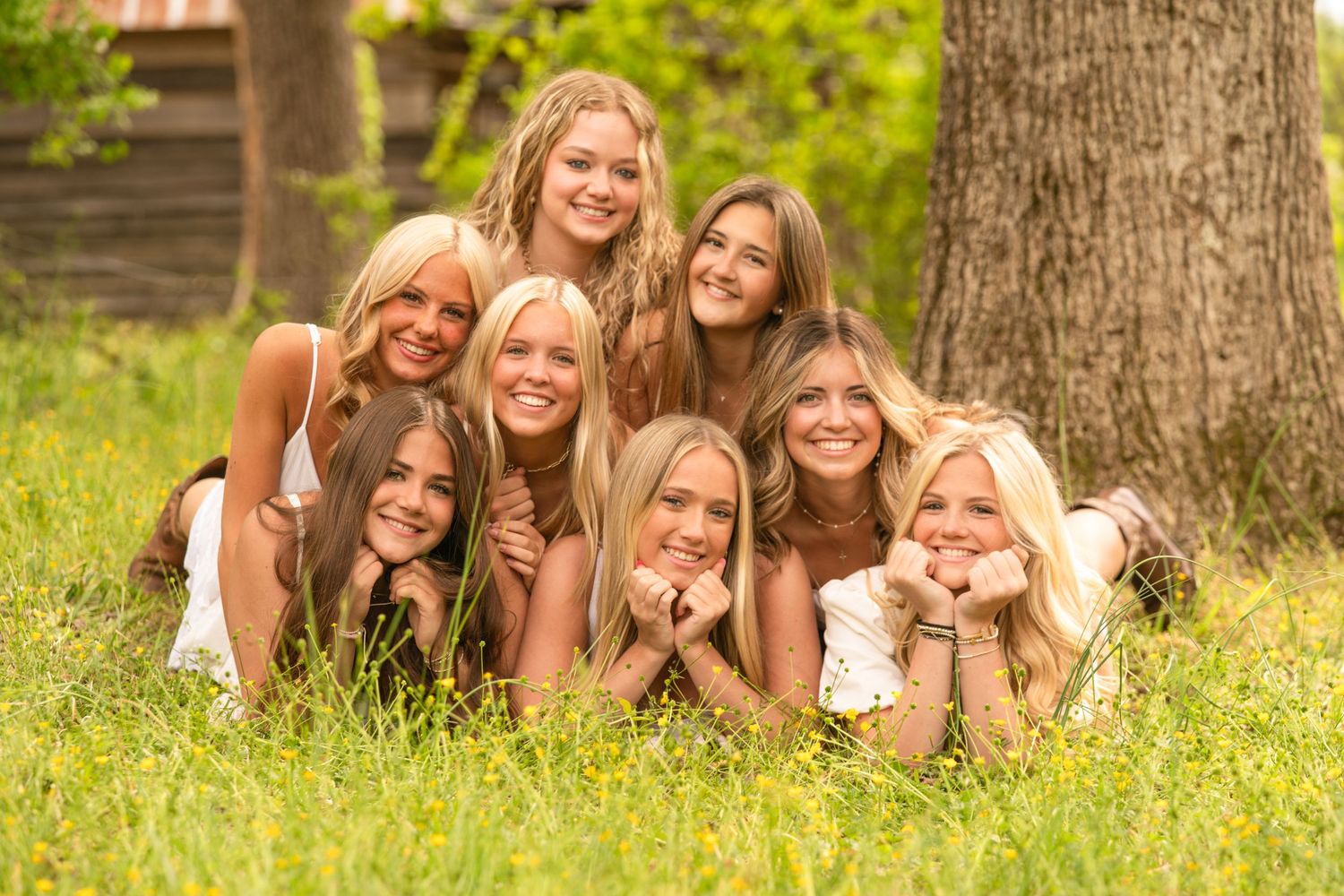 Eight smiling young women pose together in lush green grass outdoors near a large tree on a sunny day.