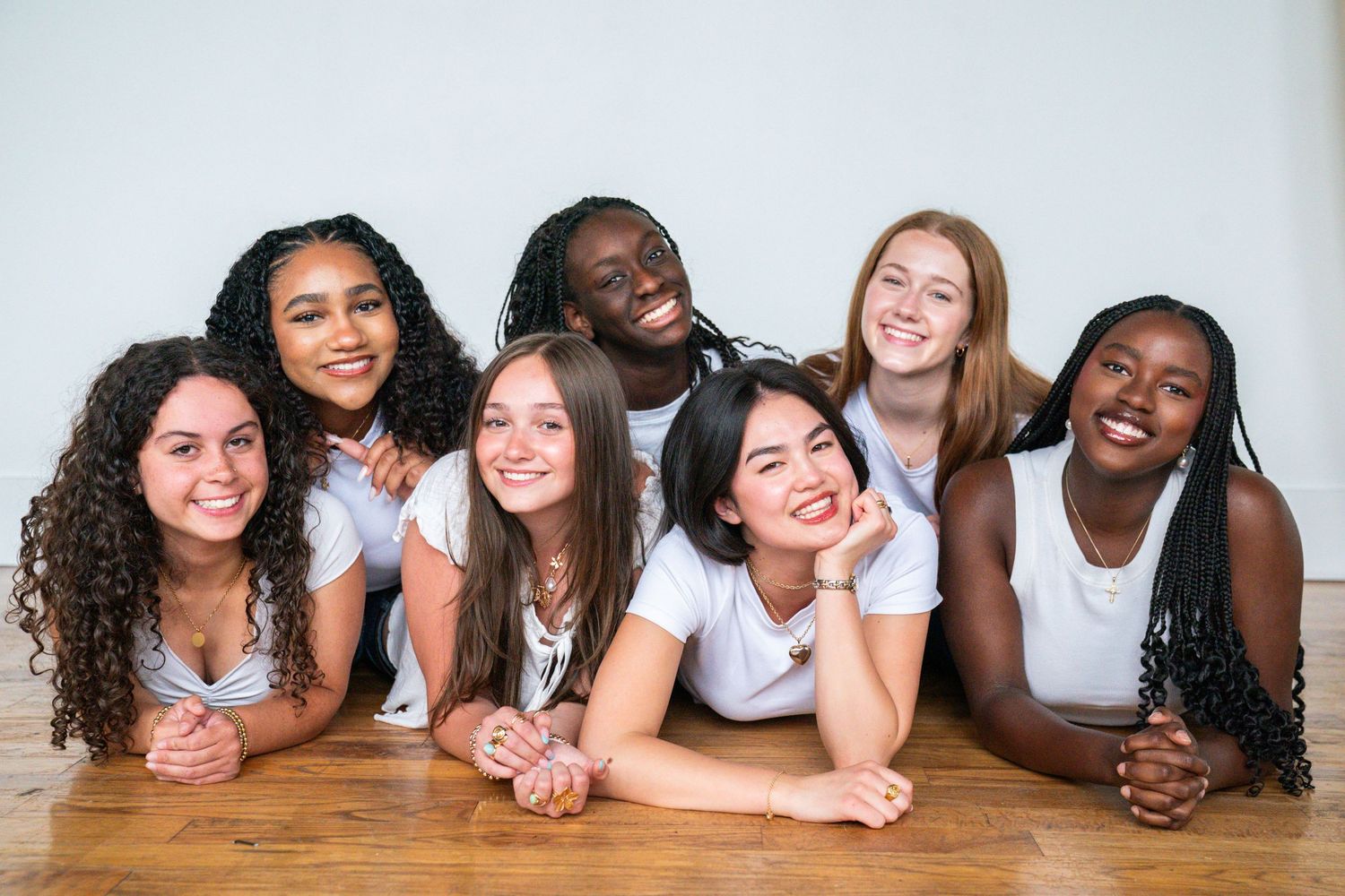 Diverse group of seven smiling young women in white tops posing together on a wooden floor against a white background.