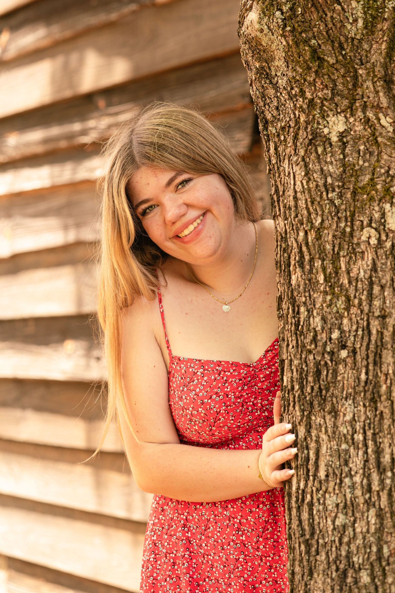 Smiling young woman in red floral dress leaning against a tree trunk near a wooden wall in warm sunlight.