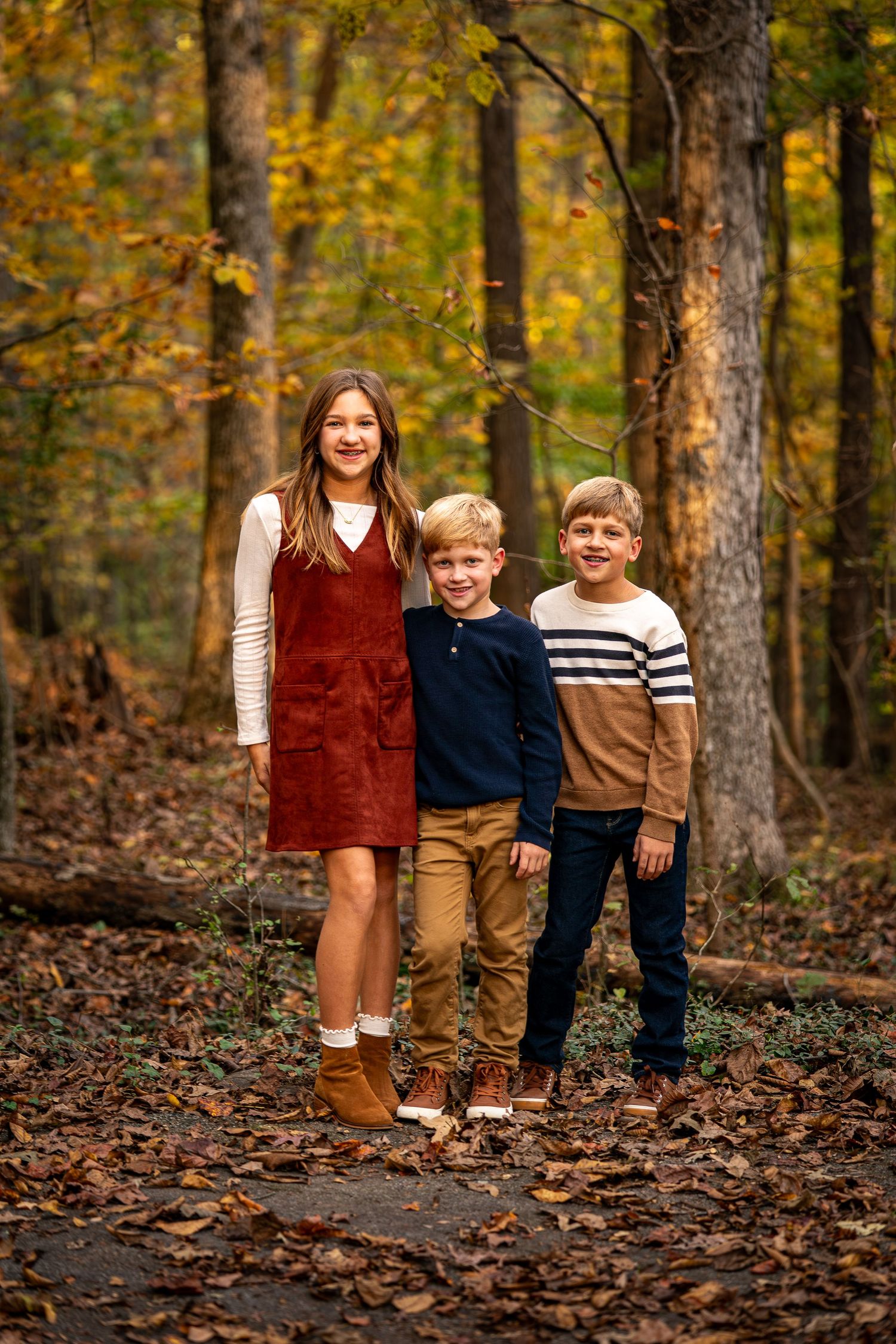 Three siblings stand together on a leaf-covered forest path during autumn.
