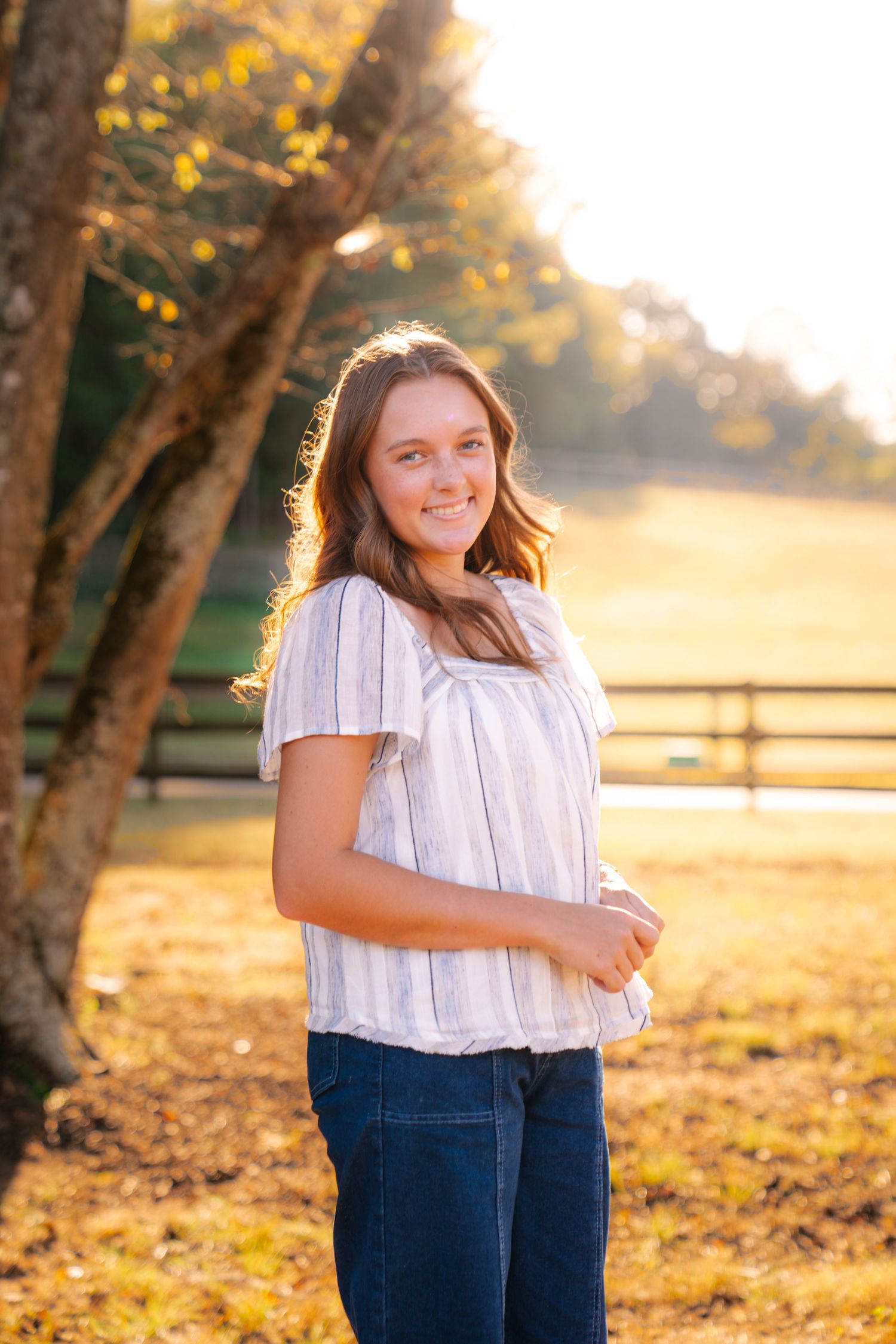 Smiling young woman with long brown hair in a striped blouse and jeans stands in a sunlit autumn field beside a tree and wooden fence.