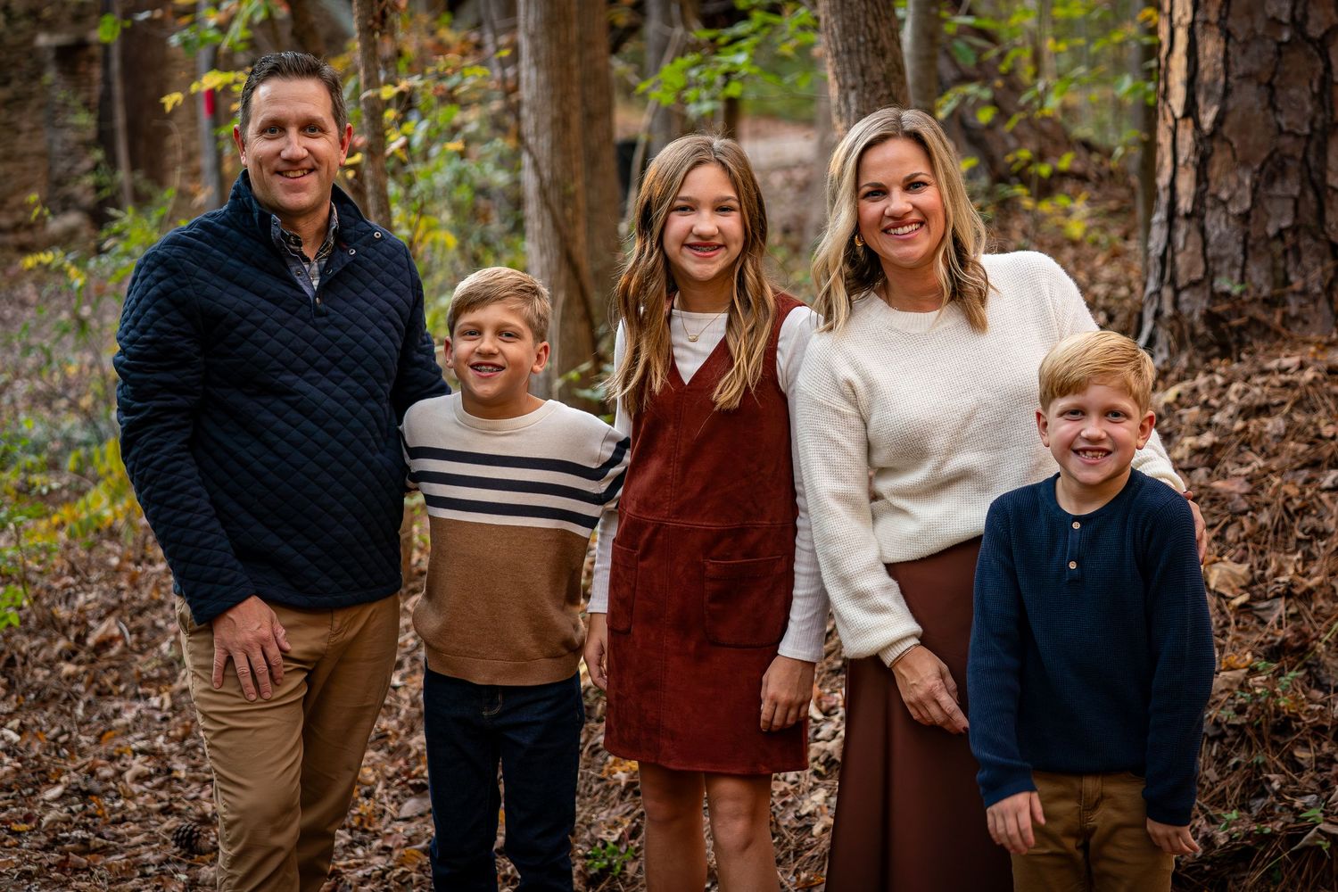 Family wearing coordinated fall outfits poses together on a wooded path with autumn foliage.