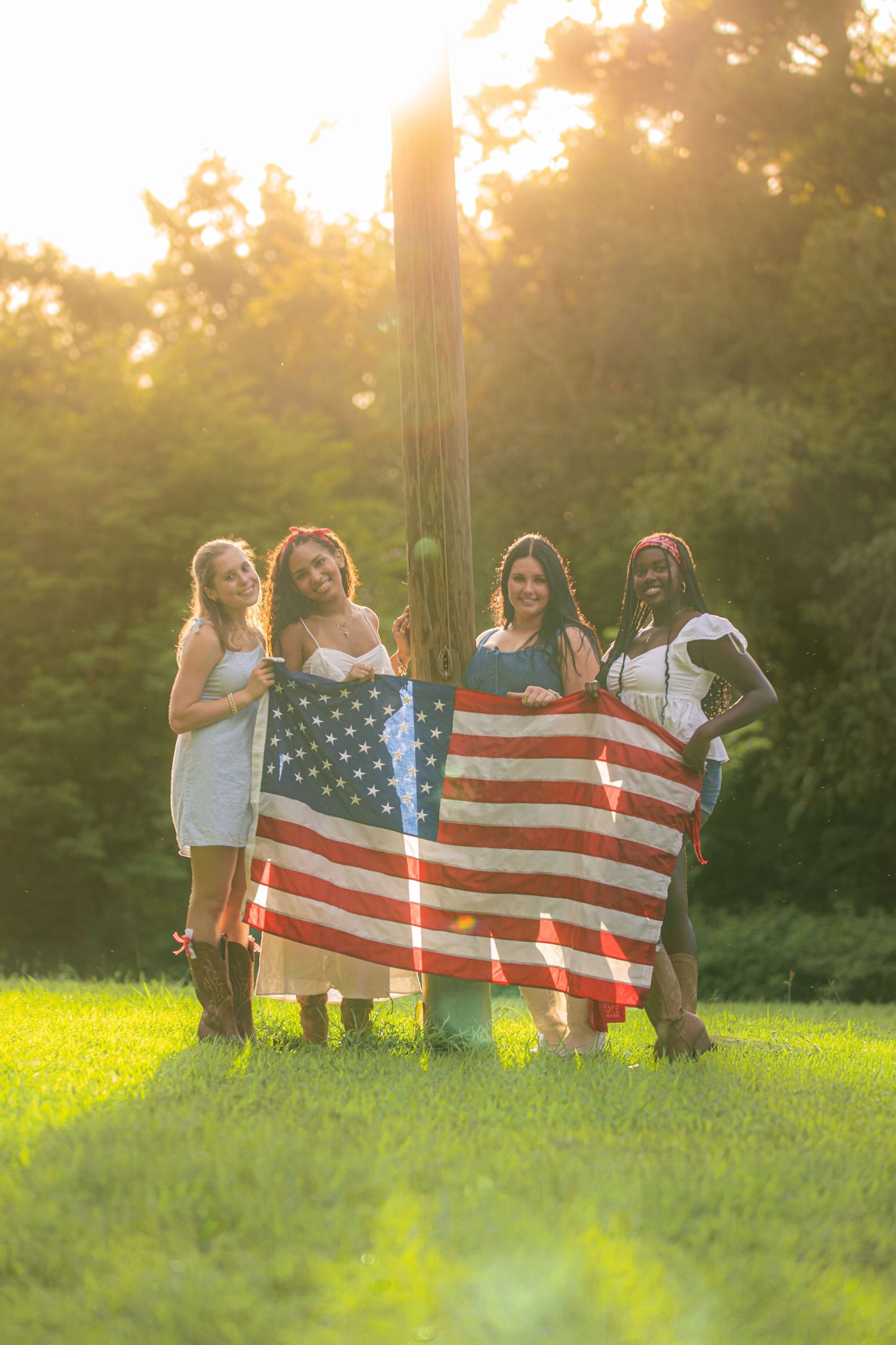 Four diverse women holding an American flag outdoors in golden sunlight, standing on green grass near trees.
