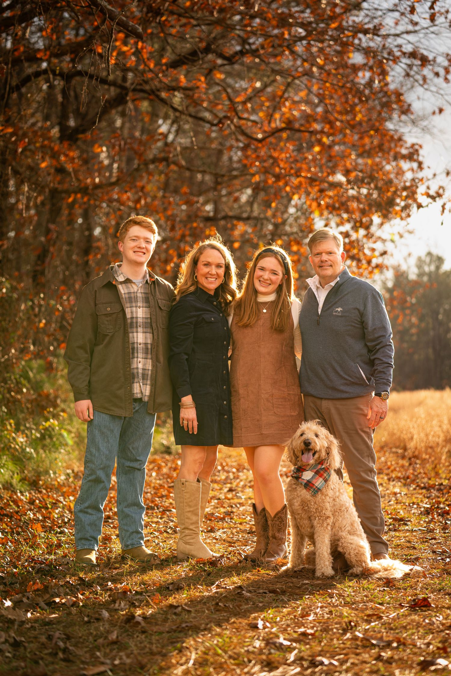 A family and their fluffy goldendoodle dog pose together on fallen autumn leaves in warm golden light.