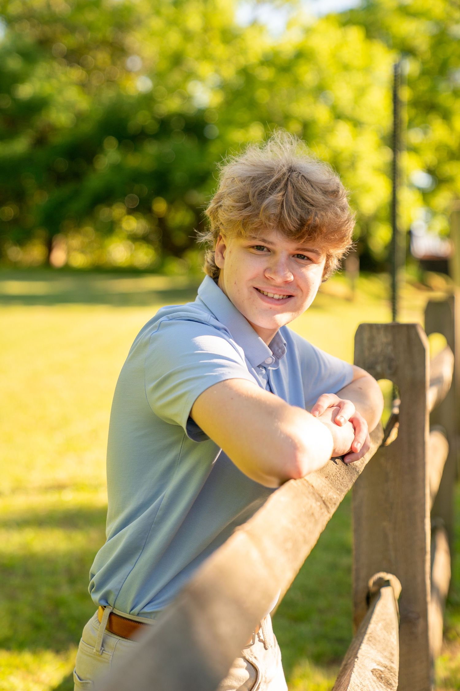 Smiling teenage boy in blue polo shirt leaning on wooden fence in sunny green park setting.