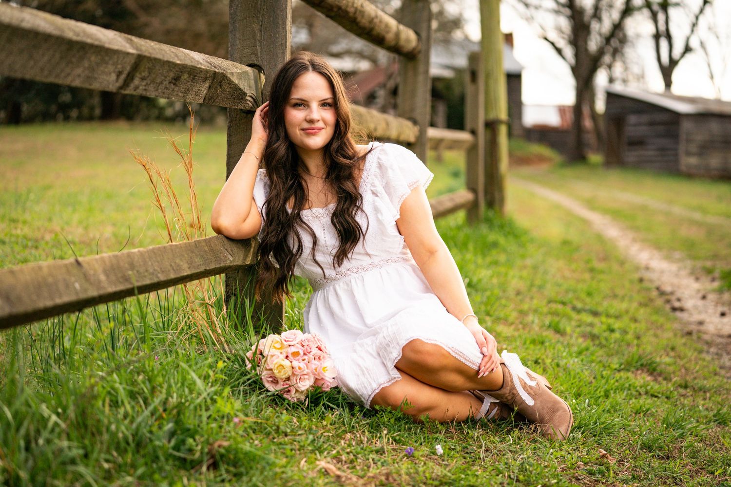 Young woman in white dress holding pink flowers, sitting by rustic wooden fence on green grass in countryside setting.
