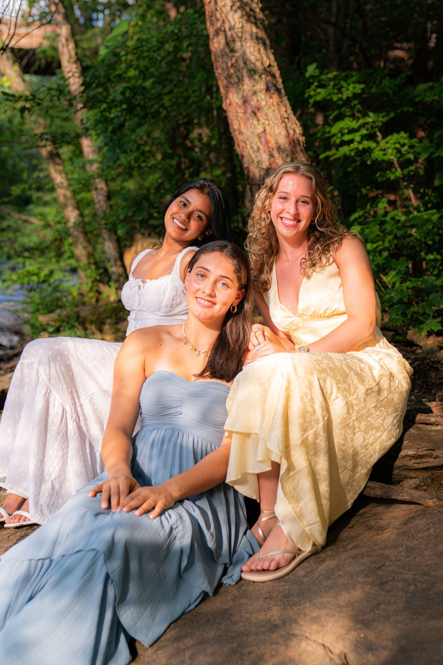 Three young women in elegant dresses pose together outdoors on rocks surrounded by lush green trees.