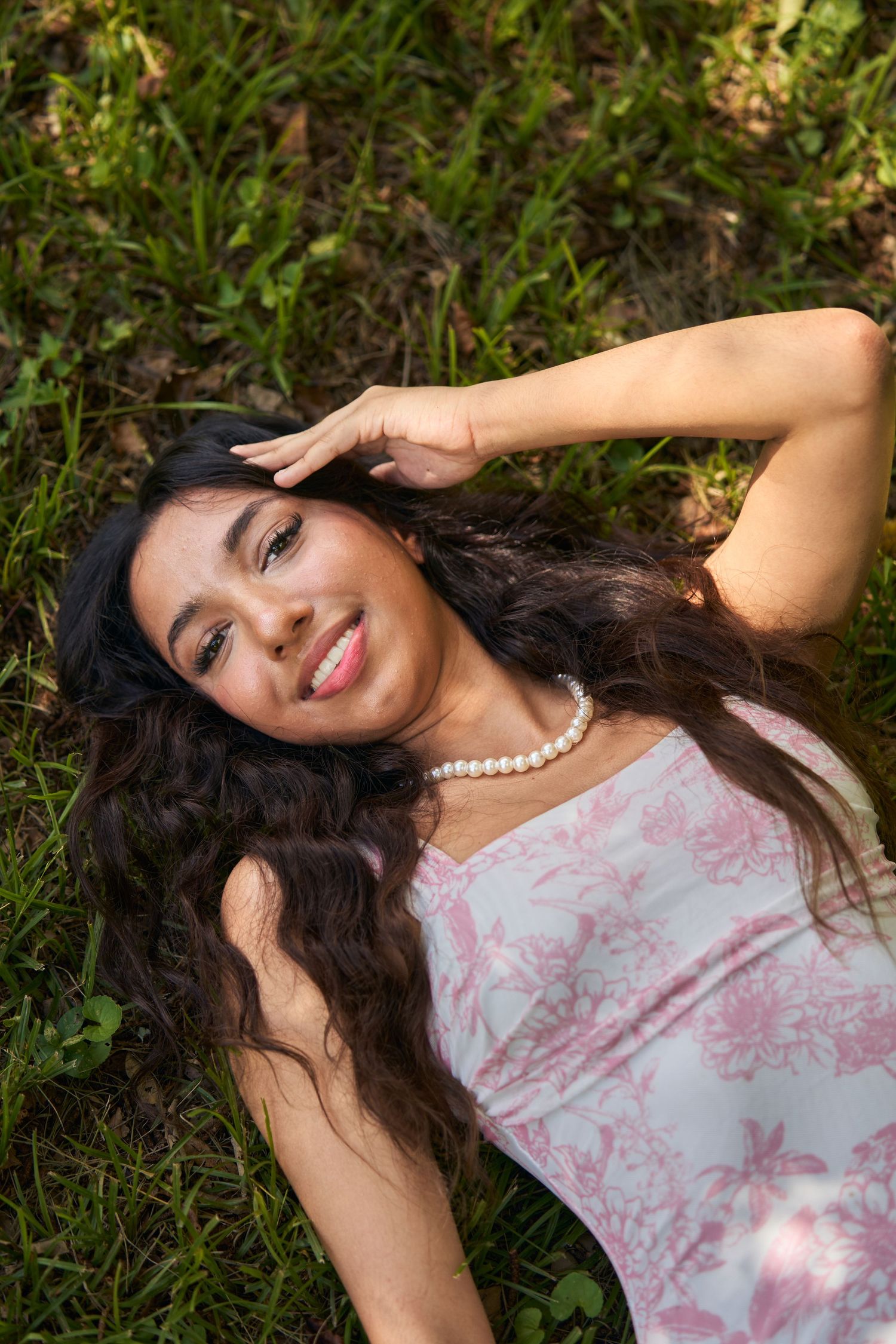 Young woman with long dark hair lying on grass, smiling, wearing a pink floral dress and pearl necklace.