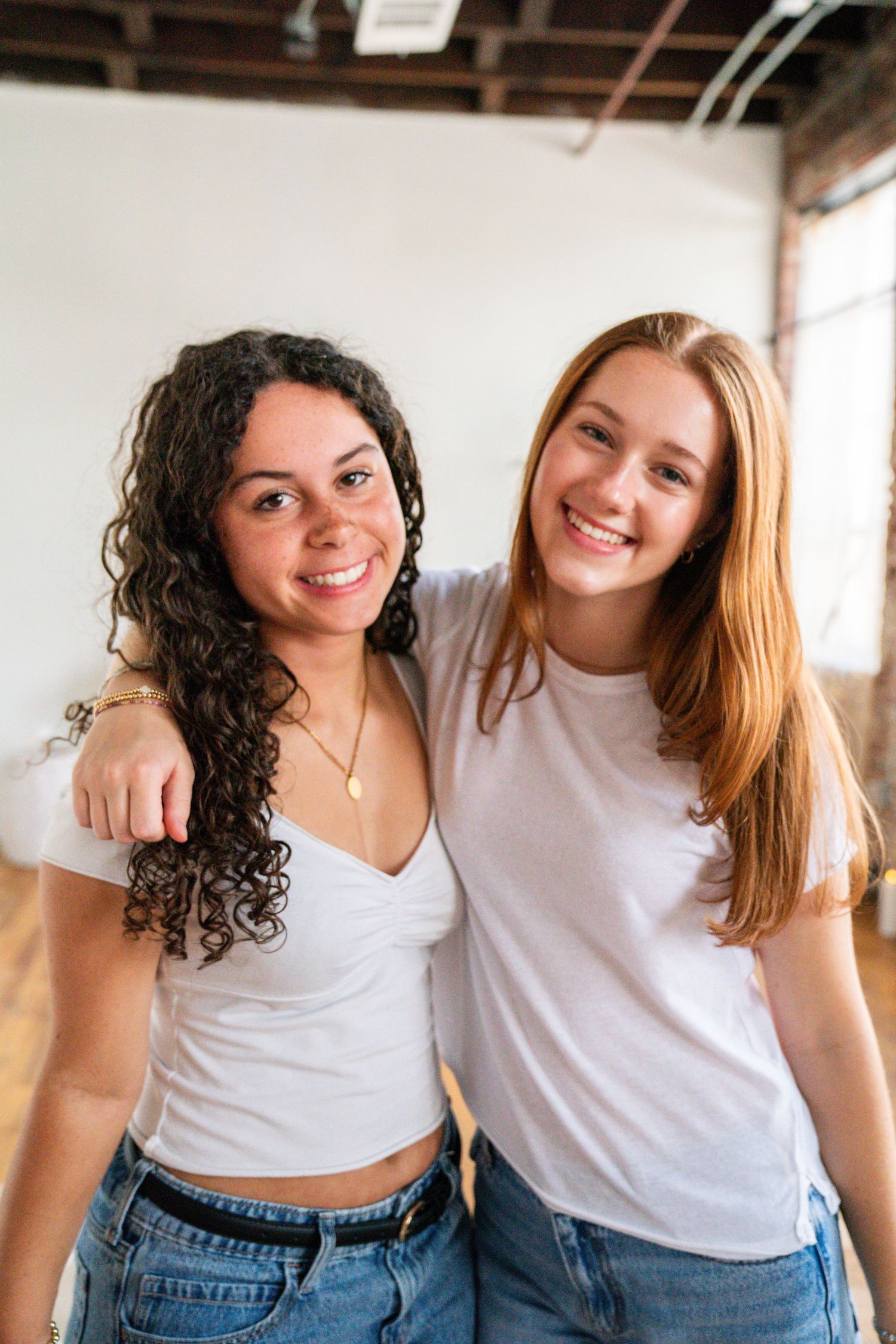 Two smiling young women in white tops and jeans posing together indoors, one with curly dark hair and one with straight blonde hair.