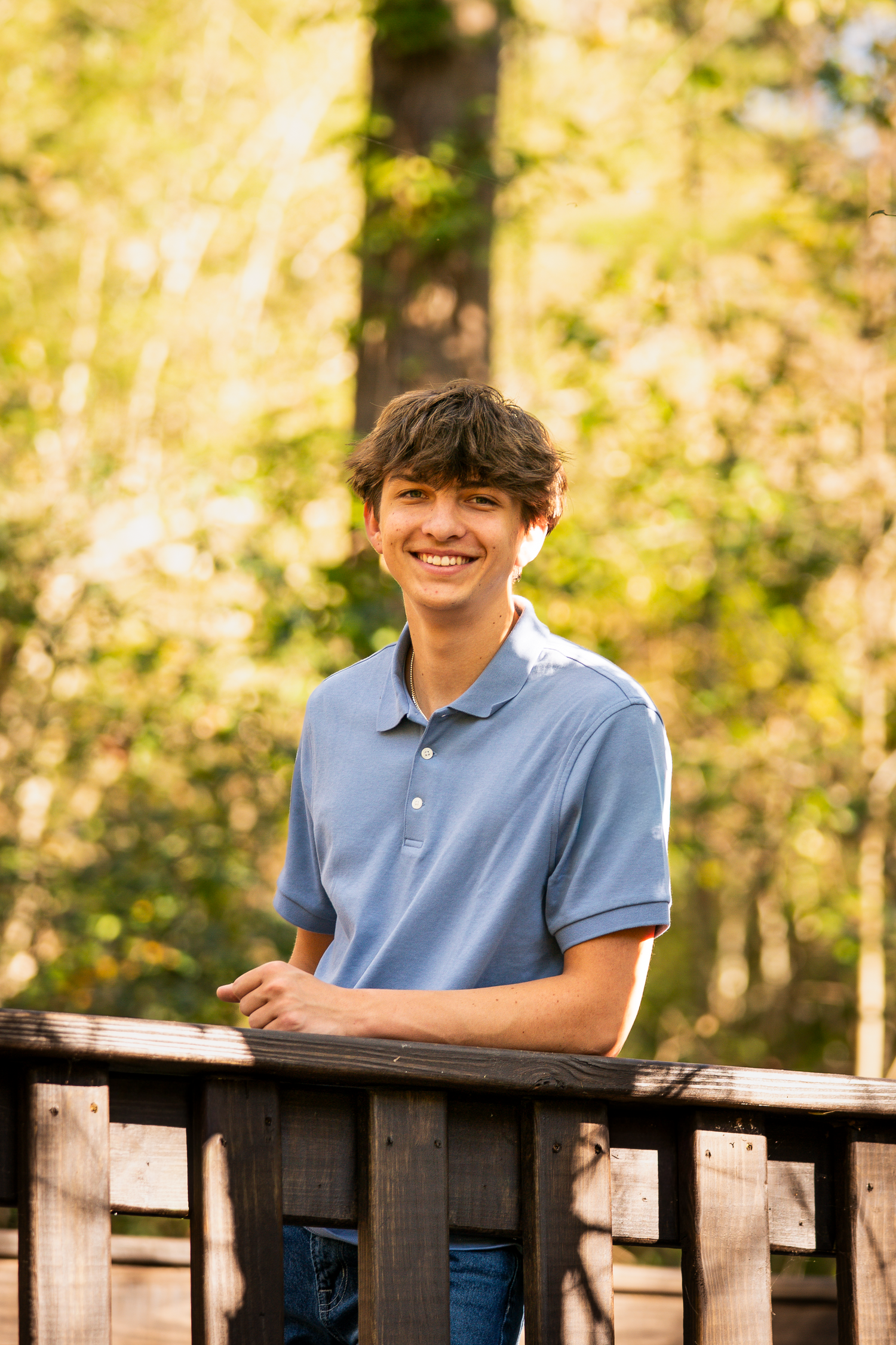 Student in blue polo shirt leaning on wooden railing with sunlit trees behind.