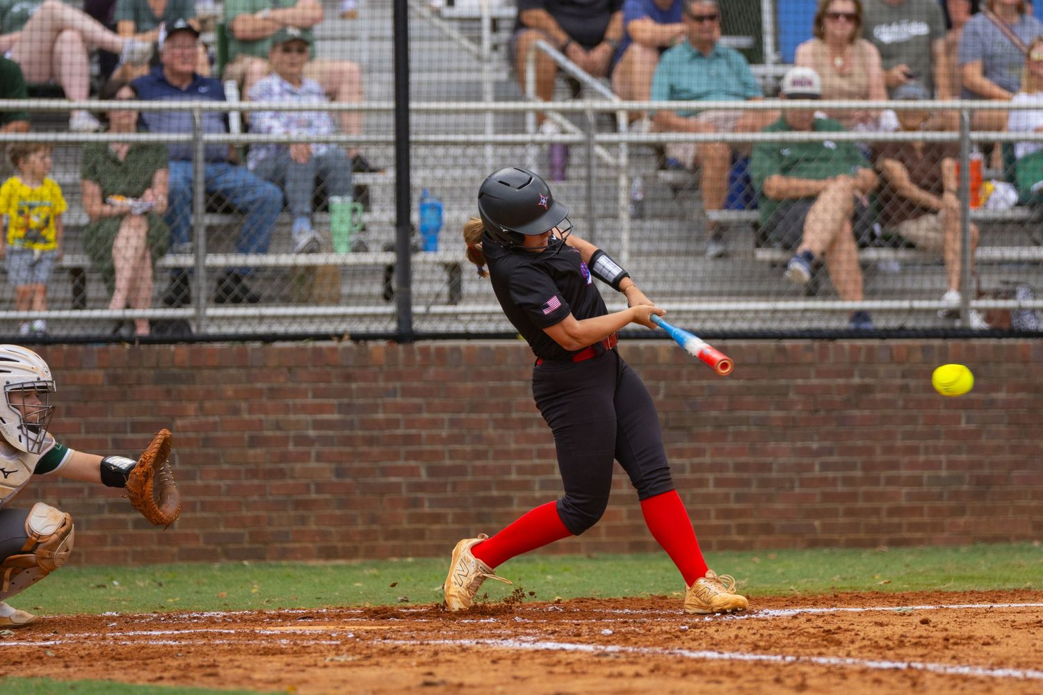 Softball batter in red and black uniform makes contact with yellow ball during competitive game with crowd in background.