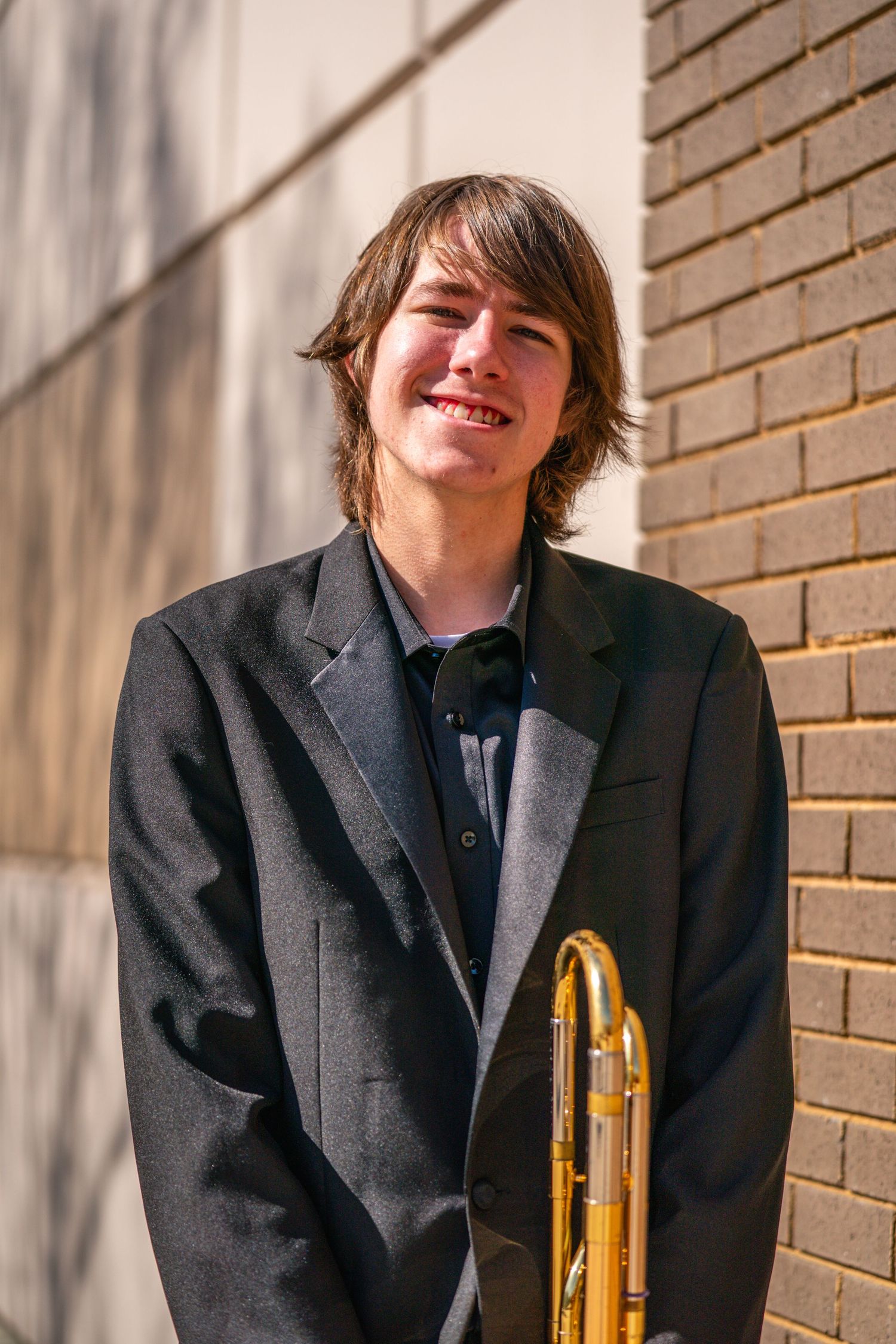 A young man with long hair wearing a black coat stands in front of a brick wall and smiles at the camera.