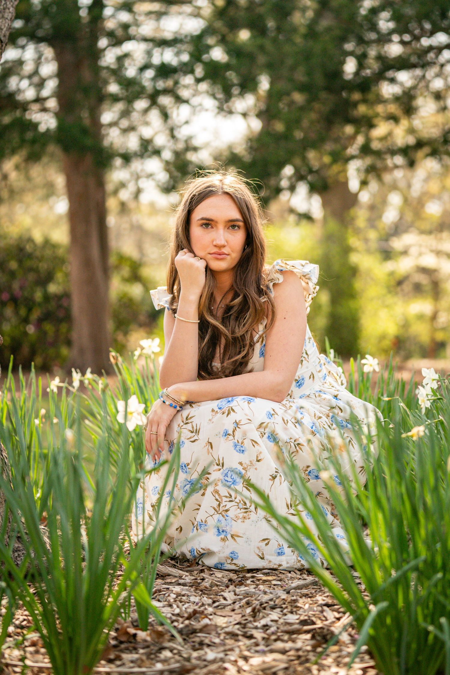 A young woman with long brown hair sits in a garden surrounded by plants and trees.