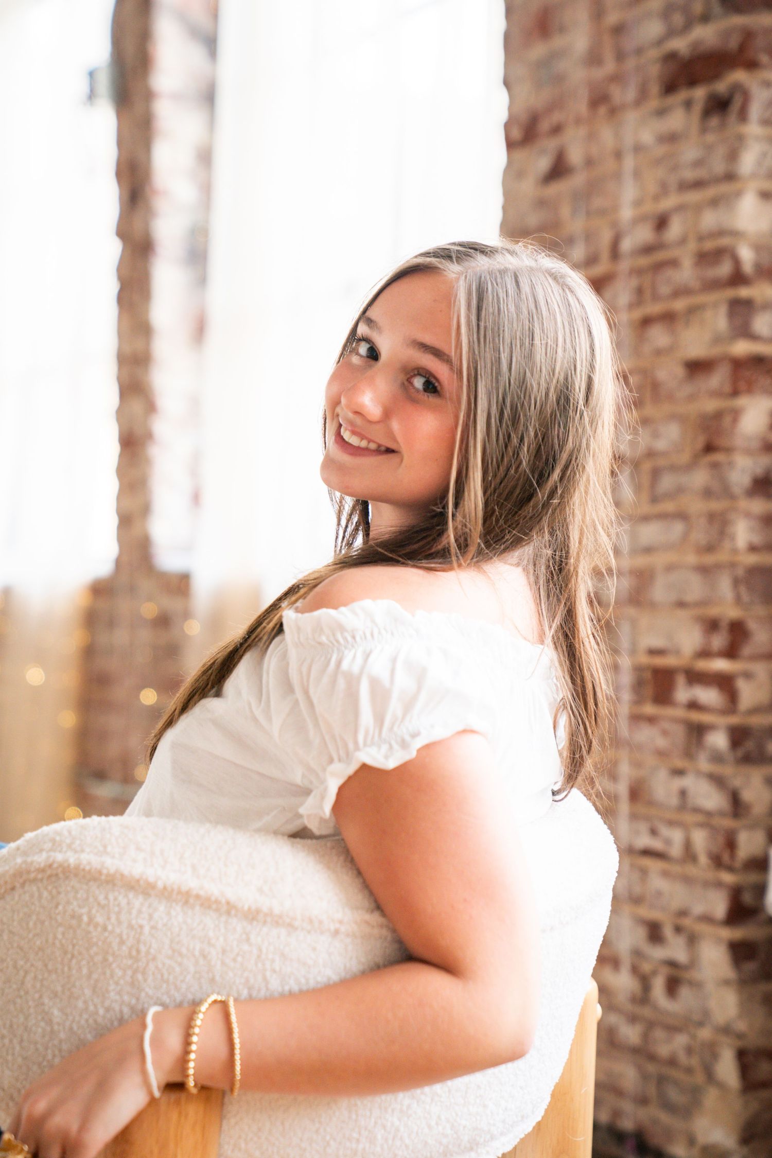 Young woman in white off-shoulder dress smiling over shoulder, seated near brick wall with natural light.