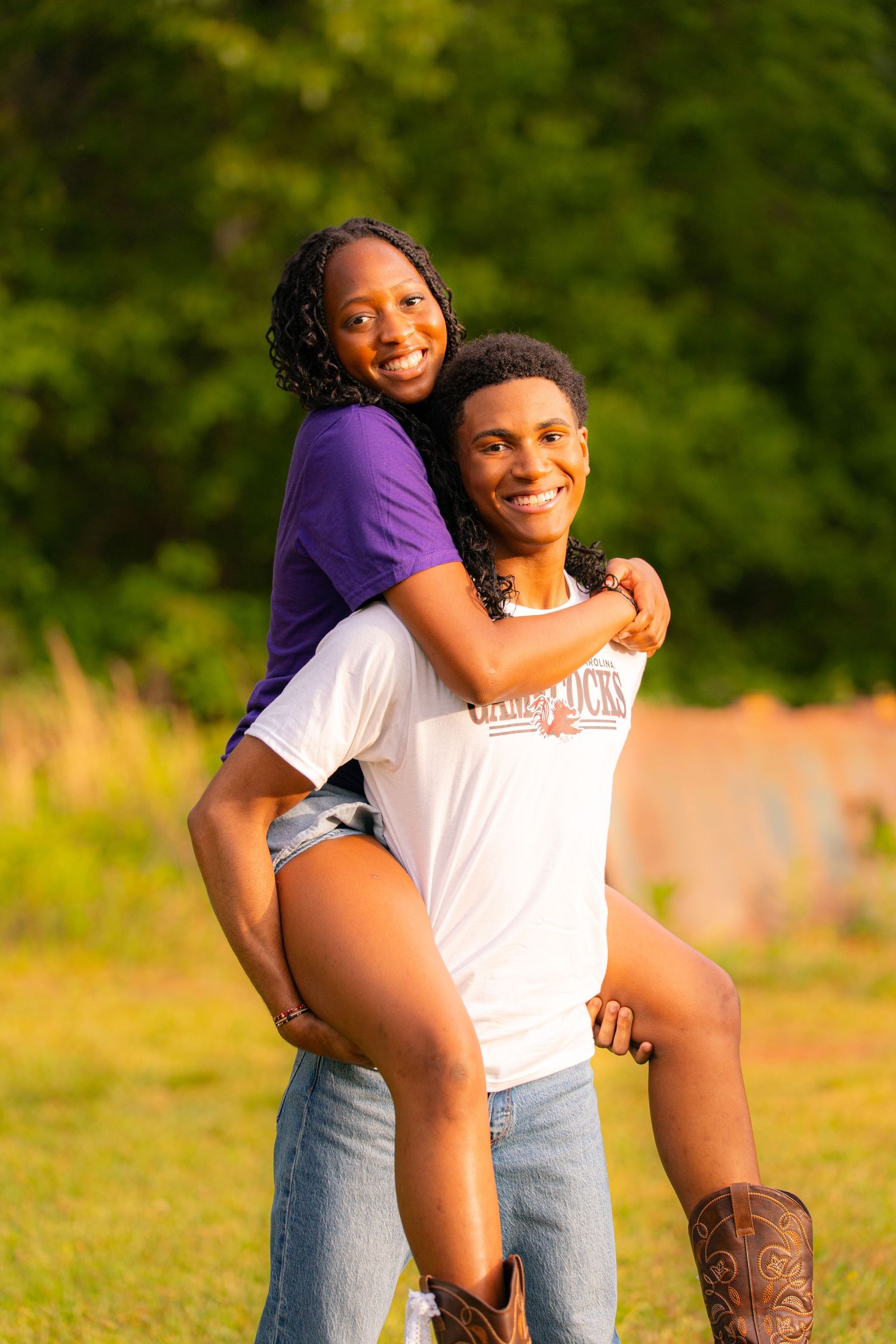A young couple smiles outdoors, man giving woman a piggyback ride in golden sunlight with green trees behind them.