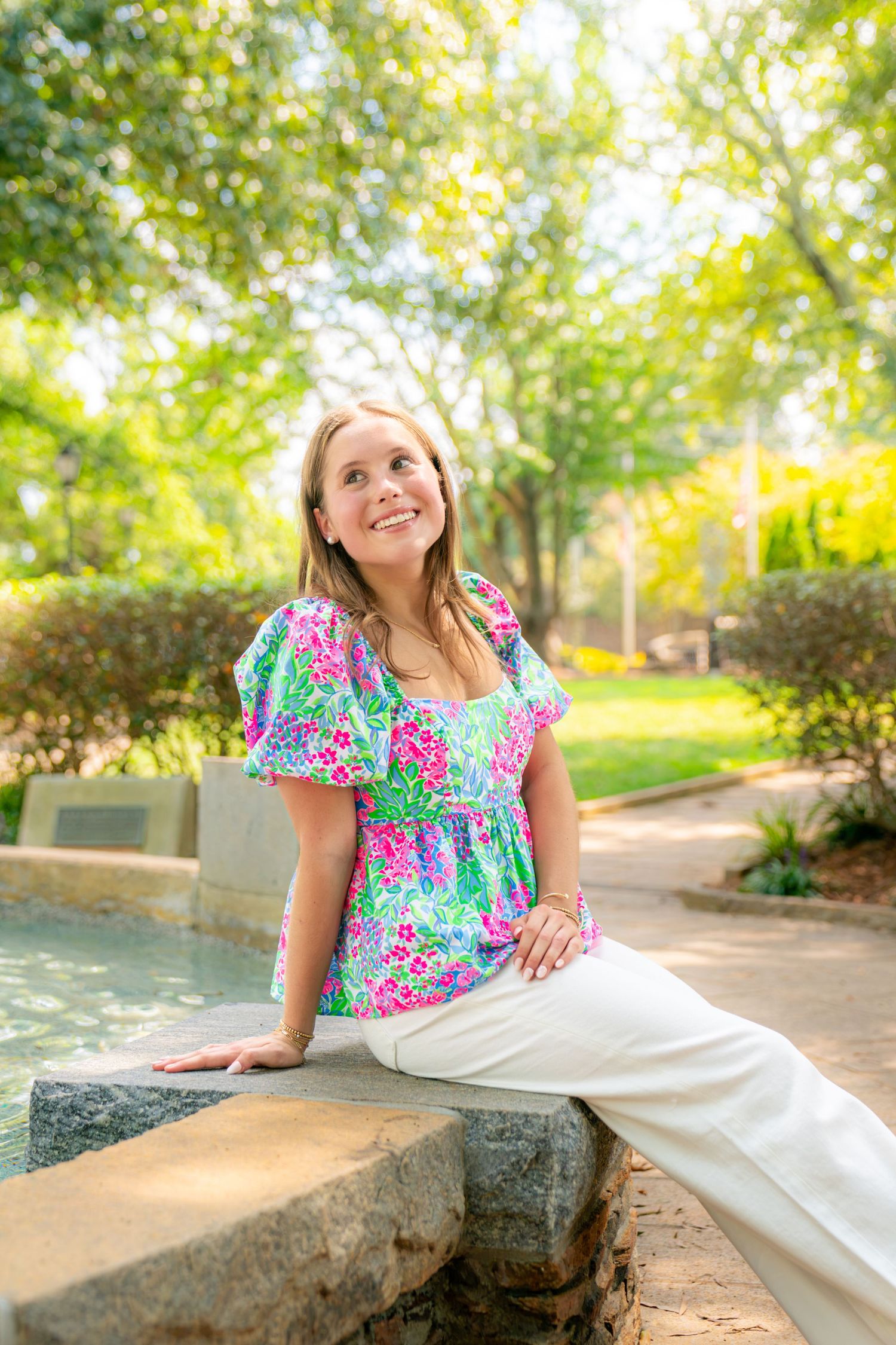 Young woman in floral top and white pants sits on stone bench, smiling in a sunny green park.