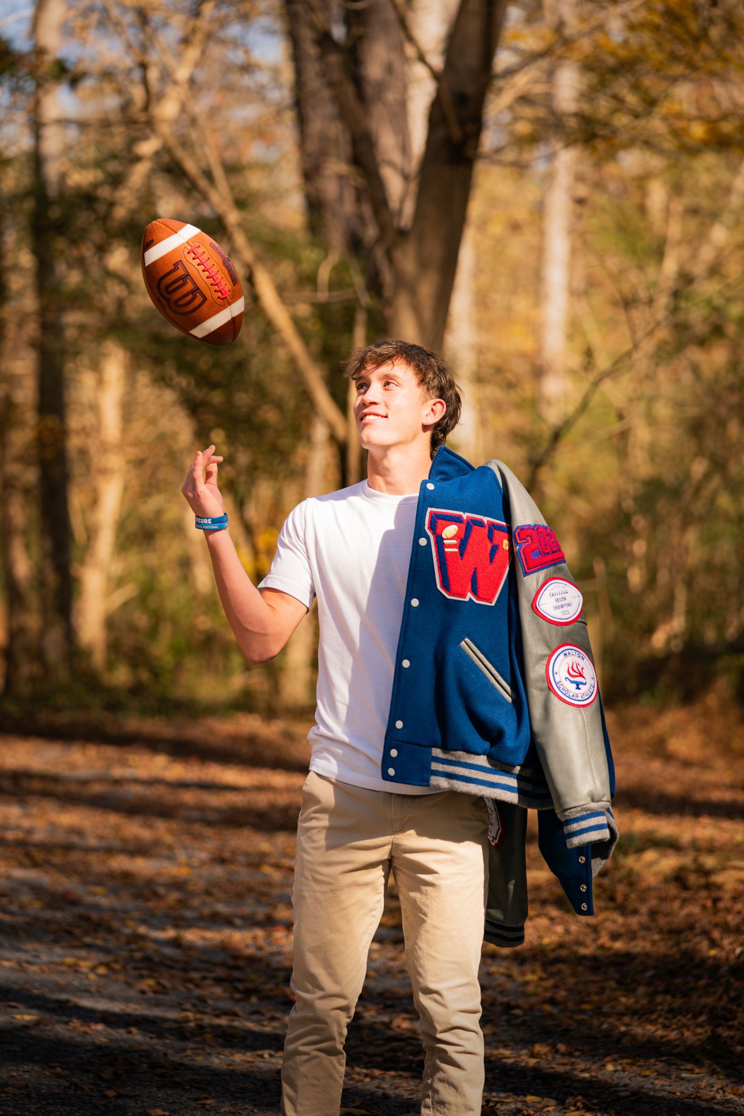 Student tosses football while wearing varsity jacket in autumn forest.