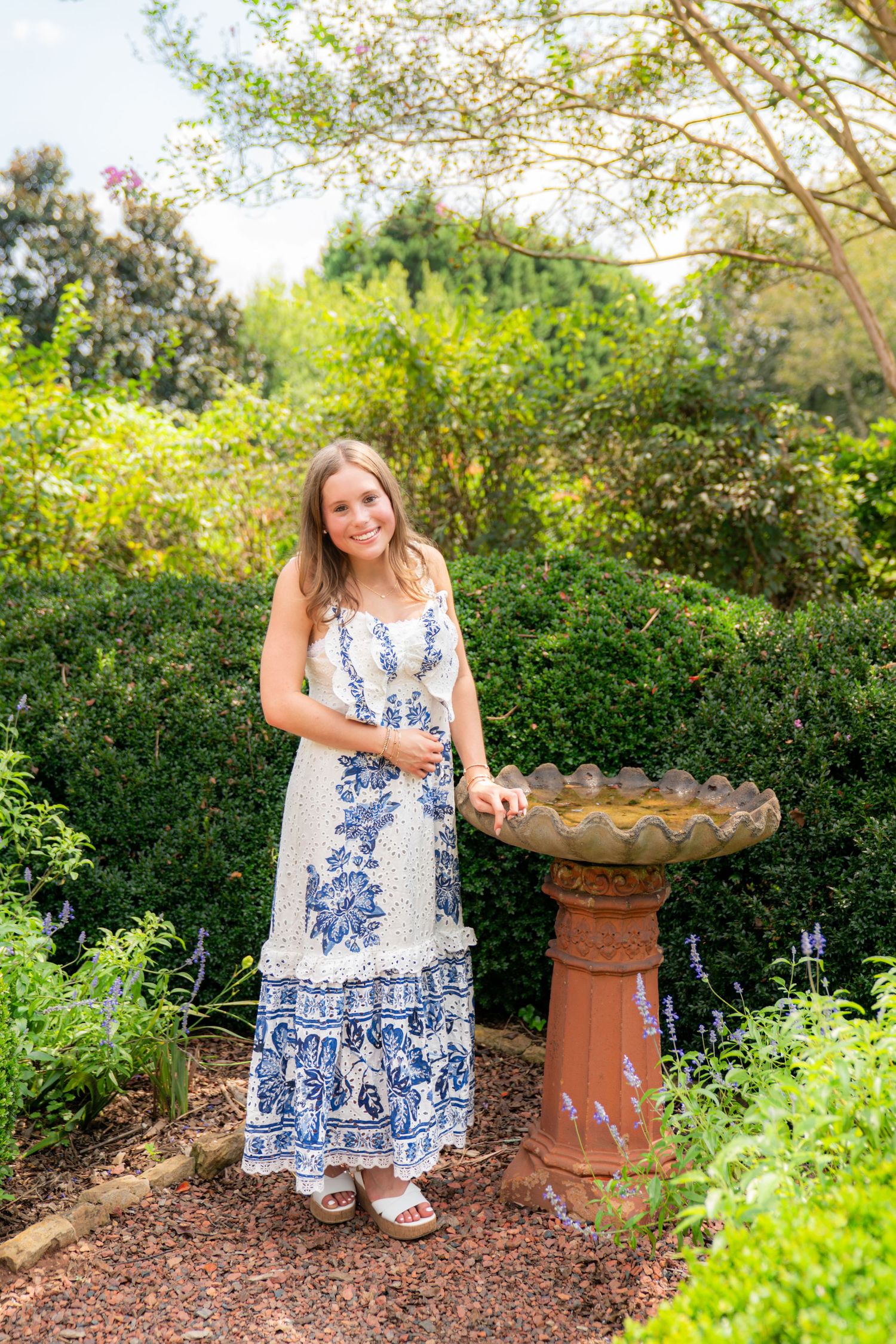 Young woman in blue floral maxi dress standing beside a birdbath in a lush green garden.