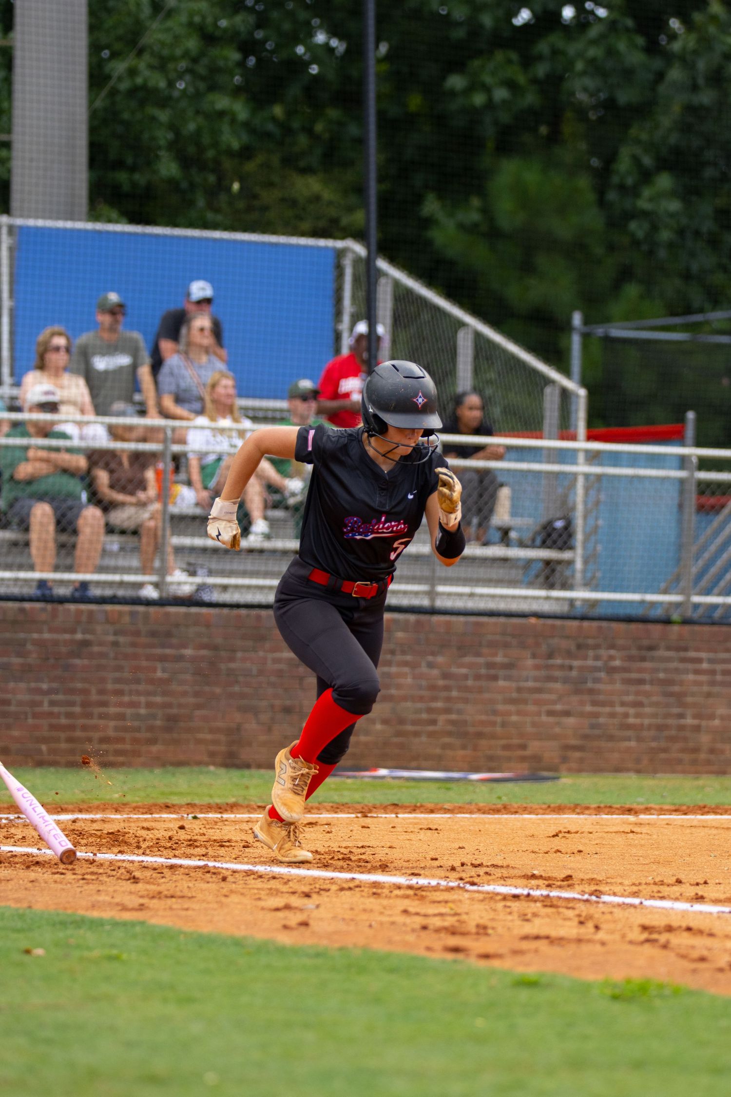 Young softball player sprints in red and black uniform on dirt field during competitive game with spectators watching.